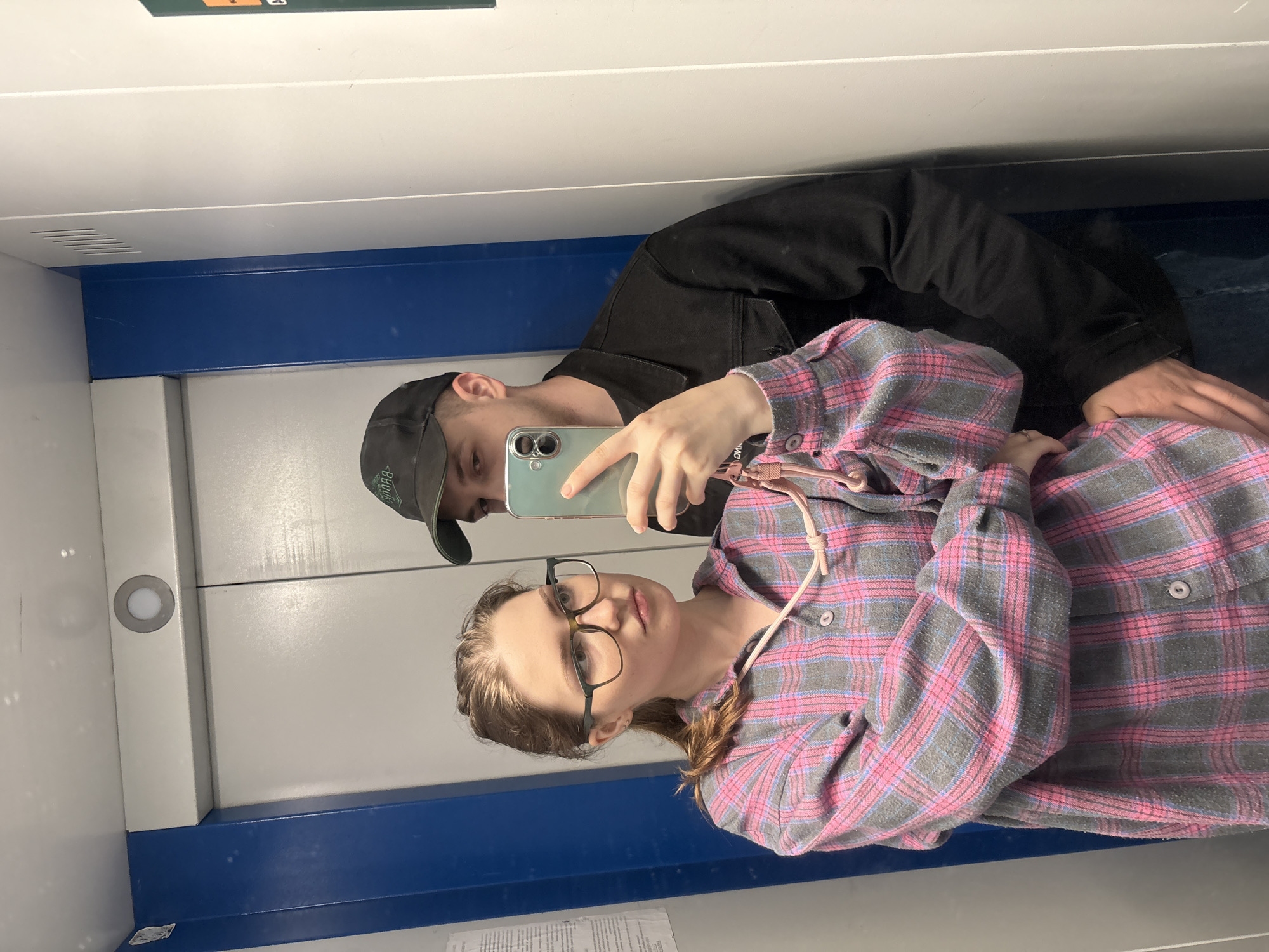 The image depicts a young couple taking a mirror selfie inside an elevator. The woman, in the foreground, is wearing glasses and a pink and grey plaid shirt. She is holding a smartphone, angled to capture the reflection. To her right, a man wearing a black baseball cap and a black jacket stands behind her. His left arm is around her waist. The elevator walls are a light grey with blue accents. The scene appears to be in an urban environment, possibly in Kirovo-Chepetsk, Russia, given the context provided.
