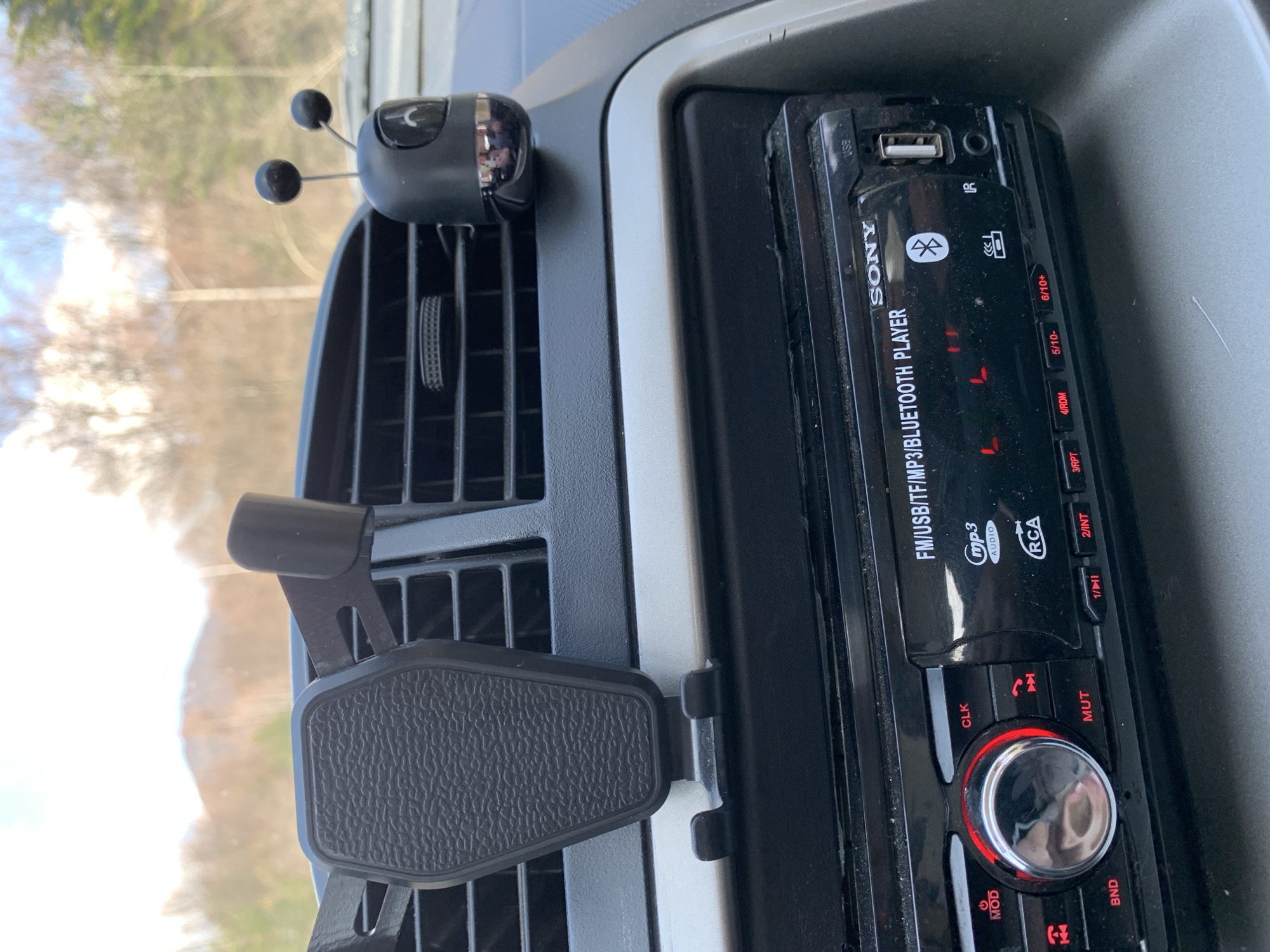 The image captures a detailed view of the interior of a car, specifically the dashboard area from a slightly tilted perspective. The main subjects are various car accessories and features.
In the center of the frame is a black Sony car stereo unit, labeled "SONY FM/USB/TF/MP3/BLUETOOTH PLAYER." It features a USB port, an AUX input, and various buttons, some of which are illuminated in red. Notable buttons include "CLK" (Clock), "MUT" (Mute), "MOD" (Mode), "BND" (Band), along with numbered preset buttons "1/||", "2/INT", "3/RPT", "4/RDM", "5/10-", and "6/10+". Symbols for Bluetooth, USB, IR, and MP3 are also visible. The stereo appears somewhat dusty.
To the left of the stereo, integrated into the air vent, is a black phone holder with a textured surface, designed to grip a smartphone. Above this holder, also attached to an air vent slat, is a small, black decorative item resembling an insect with two spherical antennae, possibly an air freshener or diffuser.
The background, visible through the car's windshield, shows a blurred outdoor scene with trees and bright sky, indicating it is daytime. The overall setting is inside a vehicle, likely in Gintsi, Bulgaria, as per the location context. No specific activity is occurring; the image is a static shot of the car's interior.