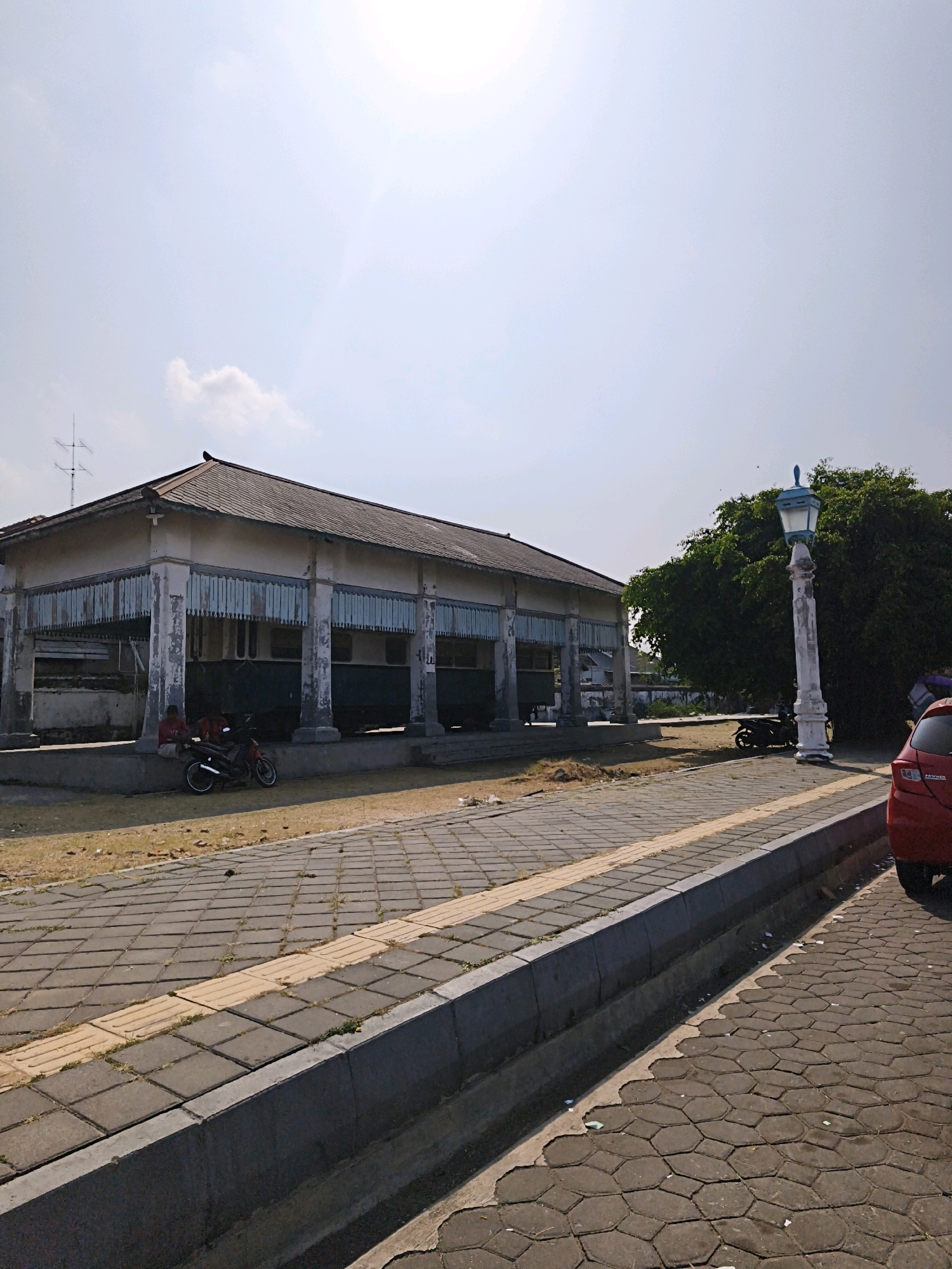 The image depicts a sunny day at the Radya Pustaka Museum in Surakarta, Indonesia. The museum's main building, a long, single-story structure with a tiled roof, is visible on the left. The building features a covered walkway supported by numerous pillars, with decorative elements hanging from the roof's edge. In front of the building, two individuals are seated on a low concrete barrier, with a red motorcycle parked beside them. A pathway made of square pavers extends in front of the building, leading towards a sidewalk with tactile paving. A decorative lamppost with a blue light fixture stands on the right side of the sidewalk. Behind the lamppost, a large, dense green tree is prominent, and a portion of a red car is visible on the far right. The ground in front of the building and under the tree is dry and covered with sparse vegetation and debris. The sky is clear and bright blue with some faint clouds. The overall scene suggests an outdoor public space in a city.