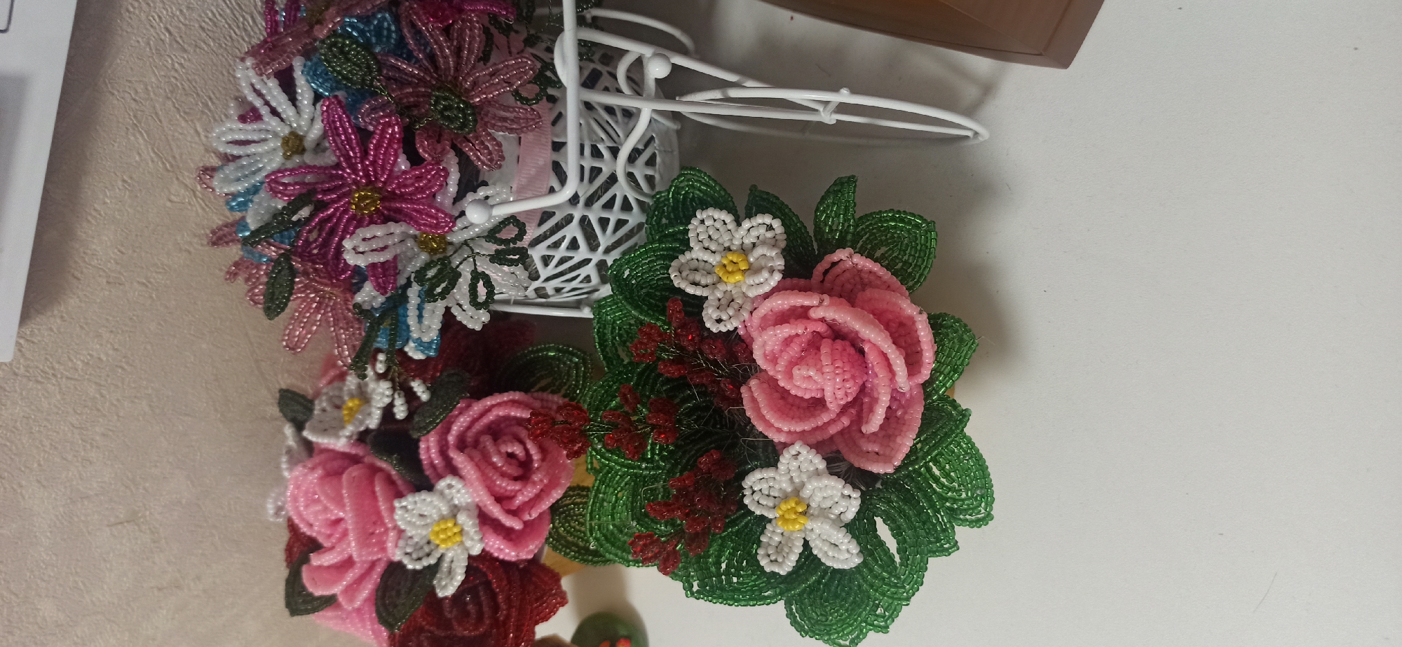 This media file features a close-up, slightly angled view of several beaded floral arrangements. The arrangement in the foreground consists of a large pink rose surrounded by green beaded leaves, with two smaller white flowers with yellow centers, and some dark red beaded accents. To the left and slightly behind this, there are more beaded flowers in shades of pink, white, and blue, many with yellow centers, cascading from what appears to be a decorative white wire basket or stand. The background is a light-colored, textured surface, possibly a wall or fabric. The lighting suggests an indoor setting with artificial light. The overall impression is of handcrafted decorative items. There are no people visible, nor any text. The location context of Asbest, Russia, does not provide any direct visual cues within the image itself.