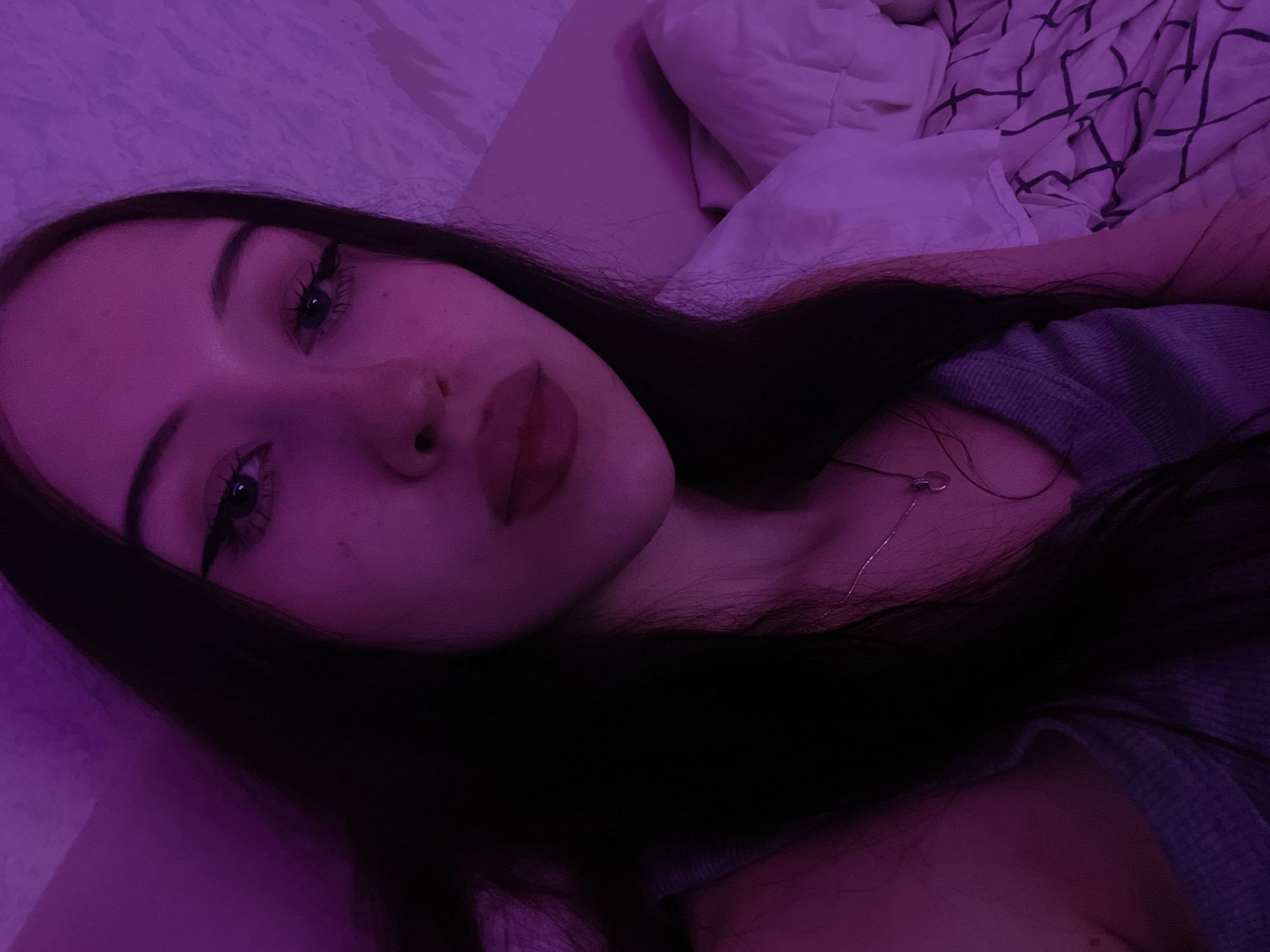 This is a close-up, selfie-style photograph of a young woman with long, dark hair, wearing a grey ribbed tank top and a delicate silver necklace with a heart-shaped pendant. She has noticeable makeup, including winged eyeliner and lipstick. The lighting is a soft purple hue, suggesting ambient or colored lighting in the room. The background shows a textured wall on the left and a bed with white bedding and a patterned duvet cover on the right.
The image is undated and unlocated, but if we were to speculate about its origin within Votkinsk, Russia, it could have been taken in a bedroom, as indicated by the presence of a bed and bedding. The overall impression is one of personal, intimate space, possibly captured at home.