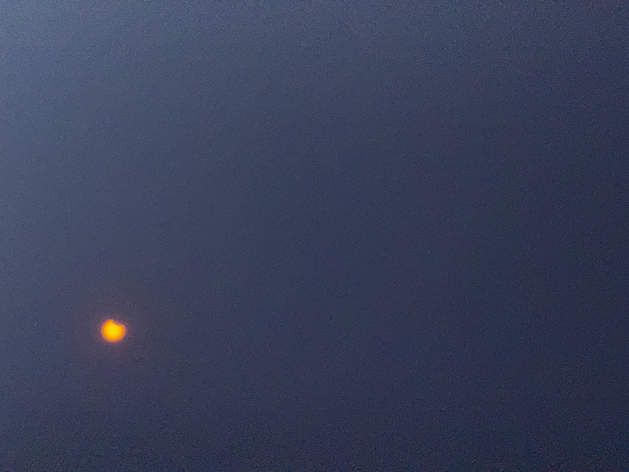 The image shows a partial solar eclipse. A crescent of bright orange light, representing the sun, is visible against a dark blue, hazy sky. The sun is partially obscured by a dark object, likely the moon, creating a distinct curved shadow. The overall impression is one of a celestial event observed through atmospheric conditions that reduce visibility and color saturation. The location is Märstetten, Switzerland.