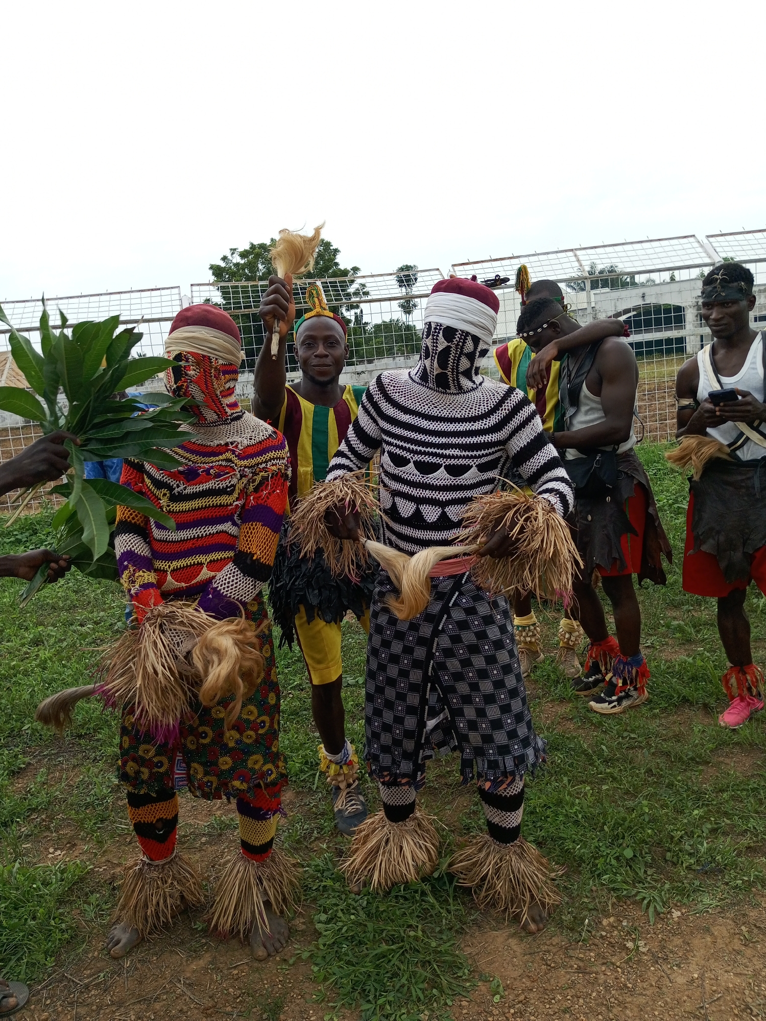The image shows a group of men standing outdoors on a grassy field. Two men in the foreground are dressed in elaborate, colorful costumes that include masks and straw-like decorations around their ankles. One man is holding green leaves. Another man is holding a tan object above his head. Three men stand in the background, casually dressed. A fence is visible in the background. The location is Takau, Nigeria.