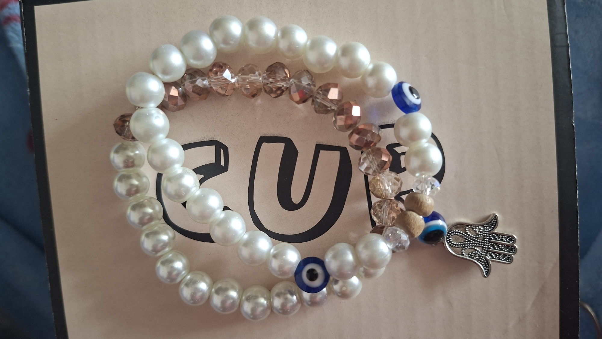 The image shows three beaded bracelets resting on a surface with a black graphic element. One bracelet is made of white pearl beads with two blue evil eye beads. Another consists of faceted, transparent bronze-colored beads. The third bracelet is a mix of pearl and bronze beads with clear crystal spacers and wooden beads, featuring an evil eye bead and a Hamsa hand charm. The background is a neutral, muted tan color.