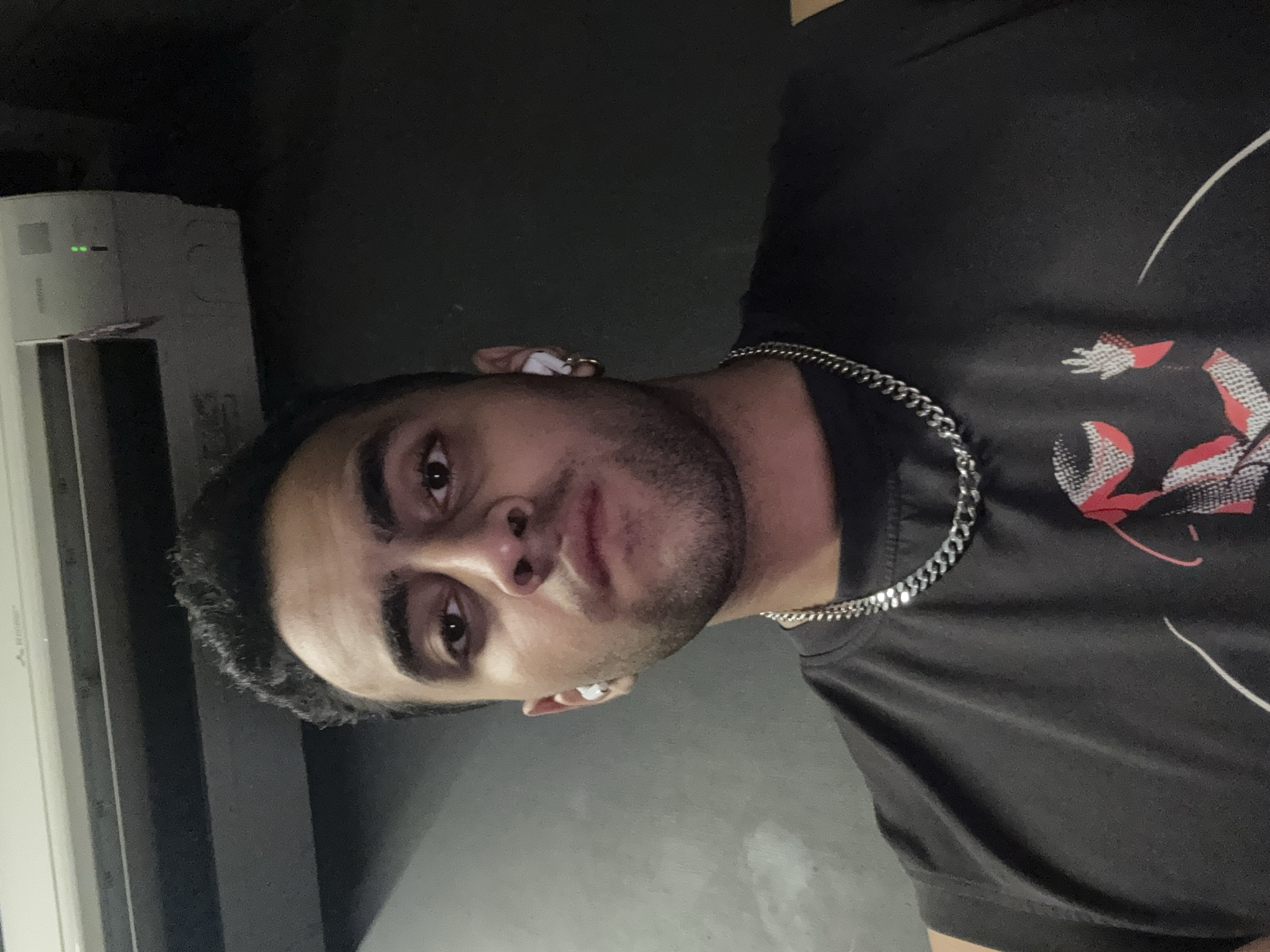 A young man with short dark hair and a trimmed beard is photographed indoors against a dark background. He is wearing a dark T-shirt with a graphic design on the right shoulder and a silver chain around his neck. He appears to be wearing earbuds. The image appears to have been taken in portrait mode, focusing on the subject with a blurred background. A white air conditioner unit is visible to the left of the man. The image provides no clear evidence that would specifically place the location in Kolkata, India.