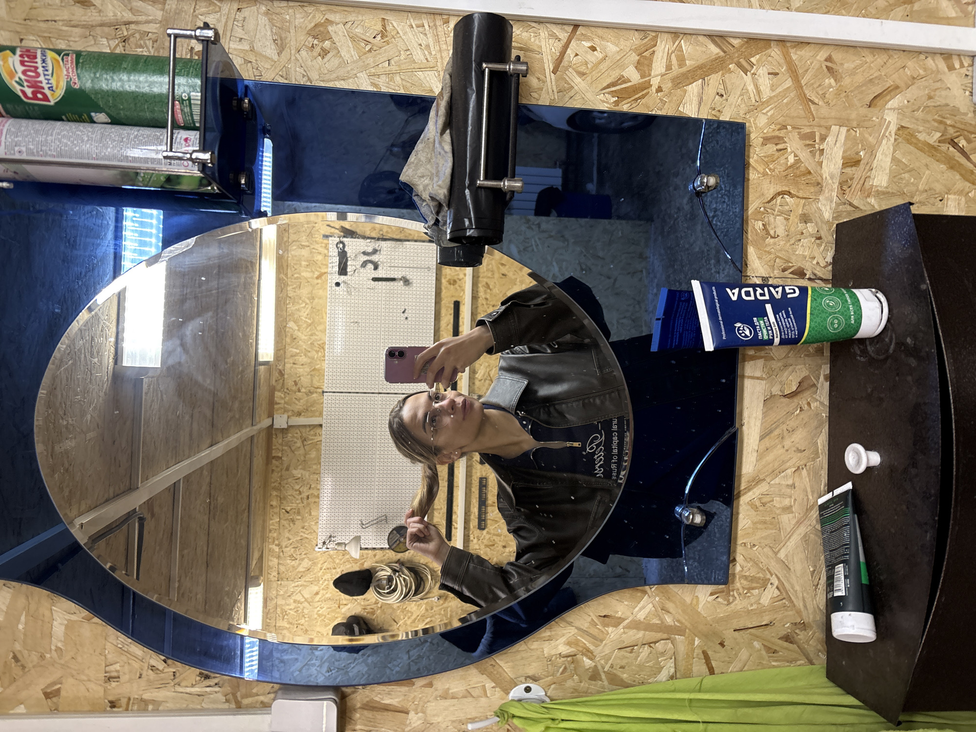 The media file, captured in Ust-Ilimsk, Russia, features a person taking a selfie reflection in a mirror. The mirror has a distinctive blue-tinted, wavy frame and is mounted on a wall made of oriented strand board (OSB), suggesting an industrial, workshop, or utility space.
The person, visible from the chest up in the reflection, appears to be female, wearing glasses and a dark jacket over a patterned top. She holds a light purple smartphone in her right hand, pointed towards the mirror for the selfie. With her left hand, she is seen pulling or adjusting a section of her hair, possibly styling it or posing for the photo.
Various objects are present in the scene. Above the mirror, a green roll of product is mounted, displaying Cyrillic text "Биолан АНТИЖИР" (Biolan Anti-Grease) and "ЧИСТАТ SROHOMий" (likely 'Чистота СУРОМЫЙ' - Purity Severe/Harsh). Another black cylindrical object is also mounted above the mirror. To the right of the main reflection, a tube of "GARDA" paste is visible, labeled with "ПАСТА ДЛЯ ОЧИСТКИ РУК И ТЕЛА" (Paste for cleaning hands and body), "Professional dermatological products", and "Nature Solo". Another black tube, possibly similar, is positioned lower on a shelf. The reflections in the mirror offer glimpses of a white pegboard and other elements of the room. The lighting is internal, suggesting either daylight filtering in or artificial illumination.