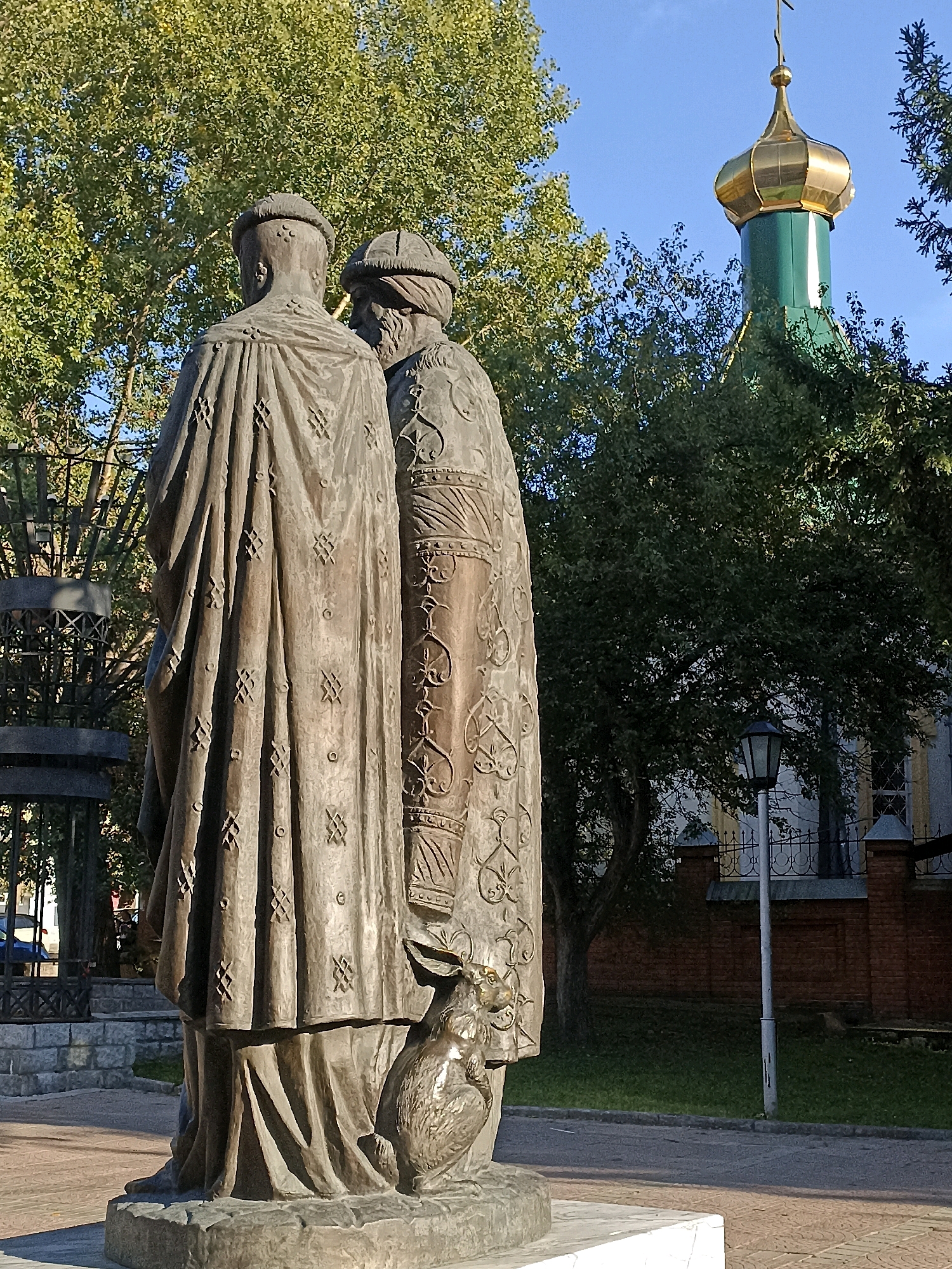 The image shows a bronze statue of two figures in historical attire, possibly royalty or religious figures, standing side-by-side. The statue is located outdoors, with a green church dome topped with a golden cupola visible in the background, partially obscured by trees. A streetlamp and a brick wall are also present. The statue is positioned on a stone pedestal, with a paved walkway in the foreground and grass behind it. The statue depicts the figures from behind, with intricate detailing on their robes. A small, animal-like figure is at the base of the statue. The sky is clear and blue.
This is the monument to Saints Peter and Paul, located in the Alexander Nevsky Cathedral square in Novosibirsk, Russia. The monument was erected in 2002 and commemorates the 10th anniversary of the restoration of the Alexander Nevsky Cathedral. The figures represent Saints Peter and Paul, who are the patrons of the cathedral.