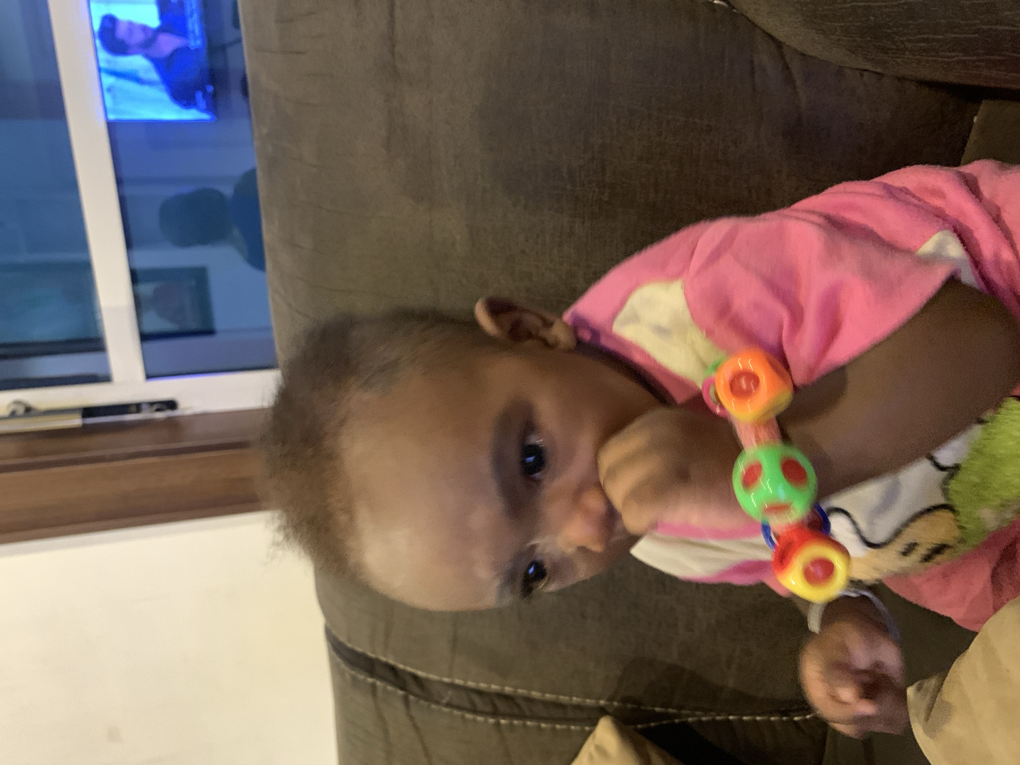 A baby lies on a brown sofa, wearing a pink outfit, and holding a colorful rattle. The baby's fingers are in their mouth. Behind the sofa is a window with a television reflected in the glass. The city of Jos, Nigeria is not discernible from the provided image.