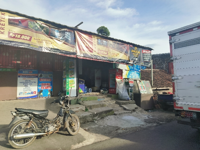 The image shows a storefront with a sign that says "KRETEK" and "Rp 18.000". The store appears to be a small grocery or convenience store, with various items displayed on shelves and signs. There is a large white truck parked in front of the store, partially visible in the right side of the image. A black motorcycle is parked in front of the store, and there are several people walking in the distance. The image was taken in Cikalong, Indonesia, with coordinates -6.732, 107.441. The scene is bright and sunny, with blue skies and some clouds visible. The ground is paved with asphalt, and there are several signs advertising goods, including a blue sign that says "ARANG ES BATU" (charcoal ice). The store facade is constructed from a mixture of concrete and metal sheeting. The truck is parked on a slightly higher elevation than the store and the motorcycle. The scene appears to be a typical commercial area in a developing city in Indonesia.
