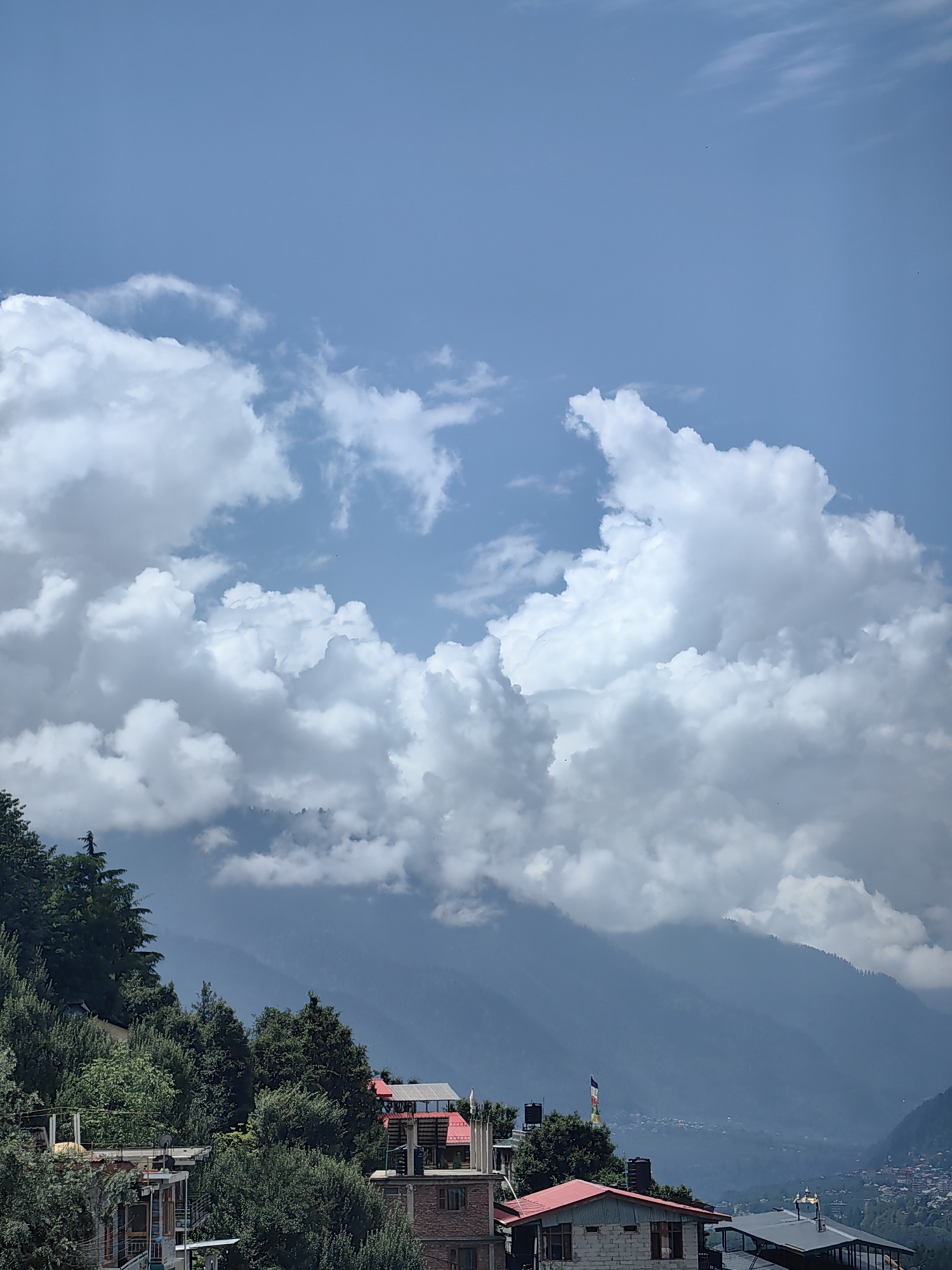 A vertical shot captures a view of Vashisht, India, showcasing a vibrant cityscape nestled amidst lush green hills under a vast expanse of sky filled with dramatic clouds. In the foreground, a hillside is dotted with multi-story buildings, their facades a mix of concrete and brick. Red-tiled roofs punctuate the scene, and several water tanks are visible. Dense foliage of trees and shrubs covers the slopes, with some buildings peeking out from behind them. The midground reveals more of the town, with its characteristic buildings and flags. Further in the distance, the landscape transforms into rolling mountains, shrouded in a soft haze. The sky above is a dynamic canvas of brilliant blue, streaked with large, fluffy white clouds that cast shadows and create a sense of depth and drama.