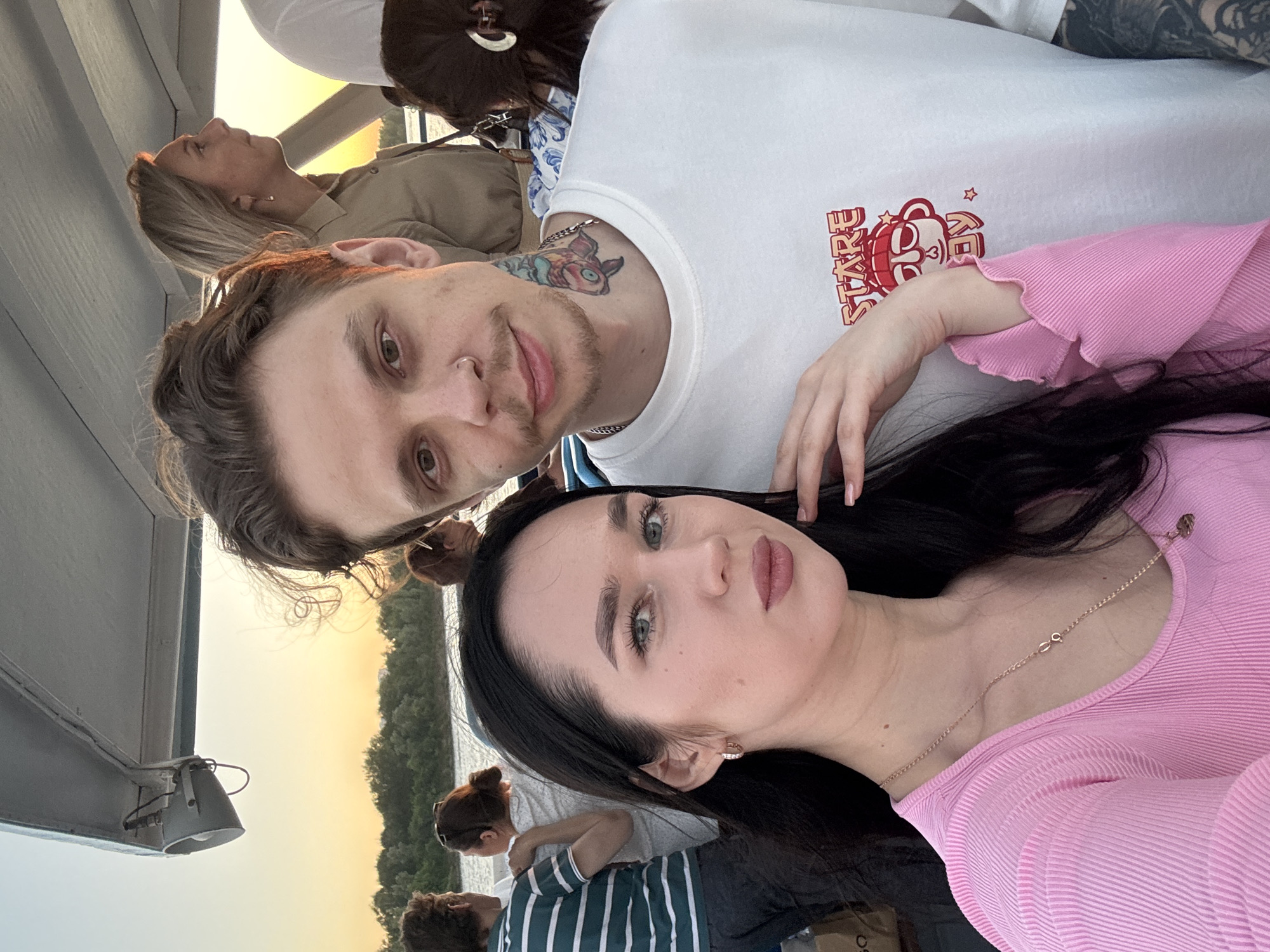 This media file shows a group of people on a boat, likely during a sunset cruise in Novosibirsk, Russia. The primary subjects are a young couple in the foreground, taking a selfie. The man has dark, curly hair and a tattoo on his neck. He is wearing a white t-shirt with red graphic text. The woman has long, dark hair and is wearing a pink, ribbed top and a necklace. They appear to be relaxed and happy.
In the background, other people are visible on the boat. One woman with blonde hair is looking up and to the side, perhaps enjoying the view. Another person, partially obscured, is wearing a striped shirt. The setting is a boat deck with a railing visible. In the background, there is a body of water and a tree-lined shore, suggesting a river or lake. The sky is a soft orange and yellow, indicating it is sunset. The overall mood is peaceful and pleasant. The text on the man's shirt is not entirely legible, but part of it appears to be "STARE."