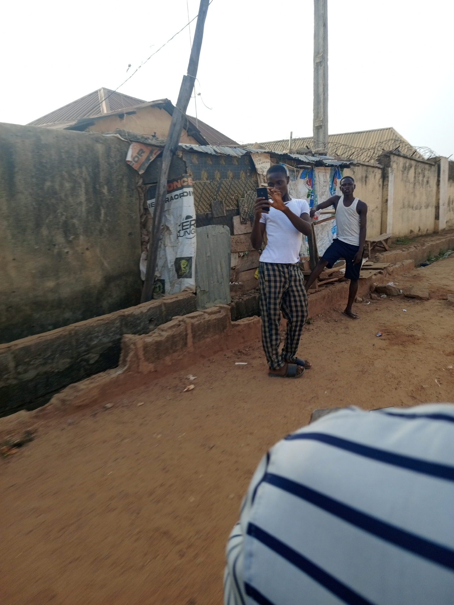 The image depicts two young men in Keffi, Nigeria. The man in the foreground wears a white t-shirt and checkered pants, holding a mobile phone as if taking a photo or video. He is standing on a dirt road. Behind him, another man in a white tank top and dark shorts leans against a dilapidated structure. This structure appears to be a makeshift dwelling or shop, with torn and faded advertisements. The background shows a mix of old buildings and a high concrete wall topped with barbed wire. The area appears to be a residential or commercial neighborhood. The foreground contains the back of a person wearing a blue and white striped shirt. The lighting suggests it's daytime, likely with overcast skies.