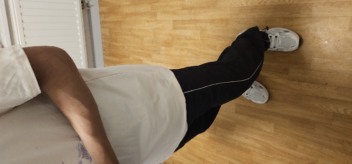 The scene depicts a person standing indoors in Kamenz - Kamjenc, Germany, viewed from a slightly elevated perspective. The subject, an individual wearing a white short-sleeved t-shirt with a faint, light purple graphic design on the left chest, black athletic pants with a single thin white stripe on each outer seam, and white athletic shoes with gray accents and white laces, occupies the central frame. The person's right arm is bent at the elbow and positioned horizontally across the midsection. The floor is constructed of light-toned wooden laminate planks. In the background, a white, horizontally-ribbed radiator is affixed to a light-colored wall, situated above a visible gray baseboard.