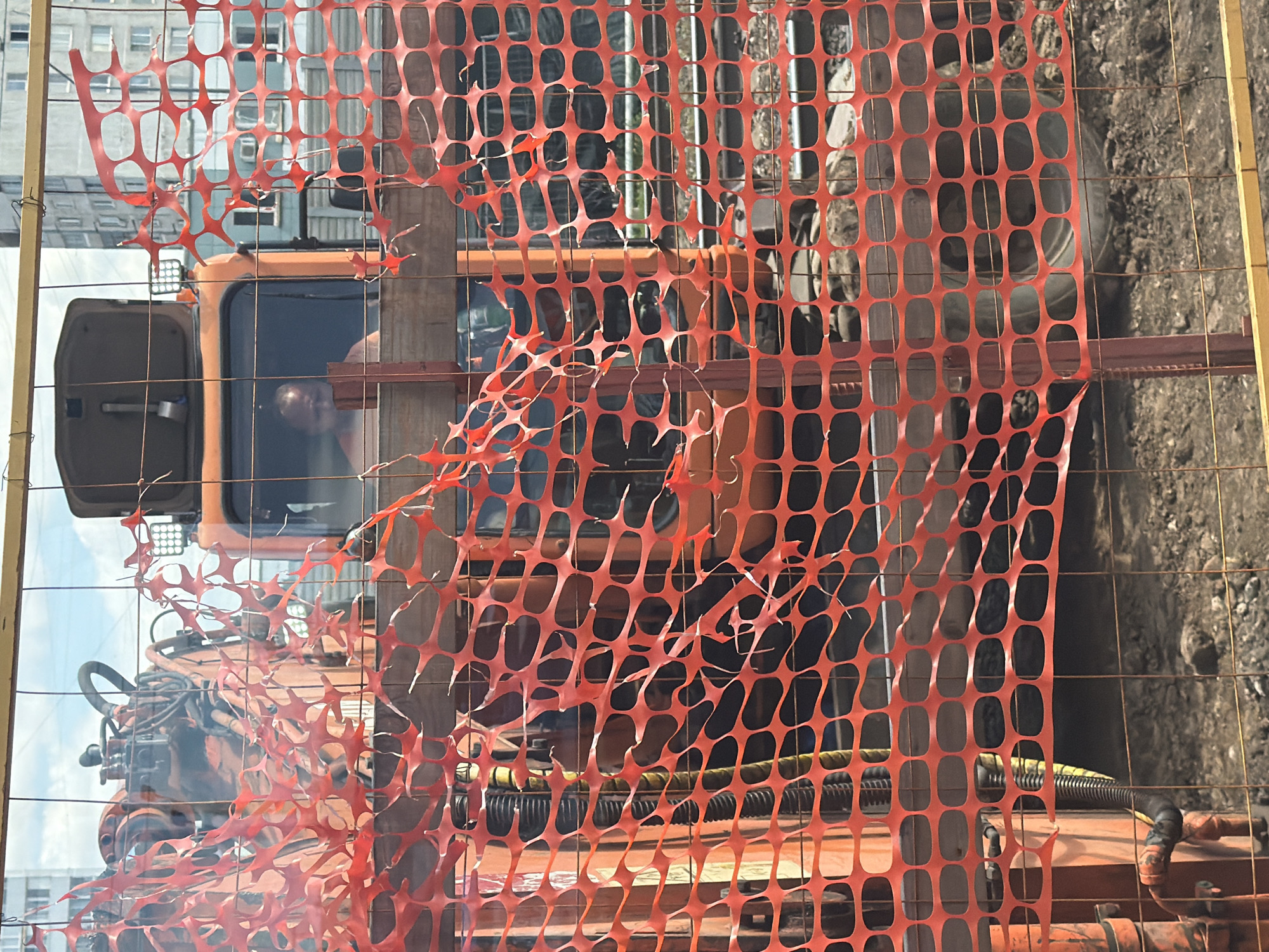 A construction site is visible behind a torn orange plastic safety fence and a wire fence. An orange excavator or construction vehicle is parked, and a man is visible through the window of the cab. The man appears to be wearing a tank top. In the background, a tall, grey apartment building is visible. The ground is dirt and gravel. The sky is cloudy.