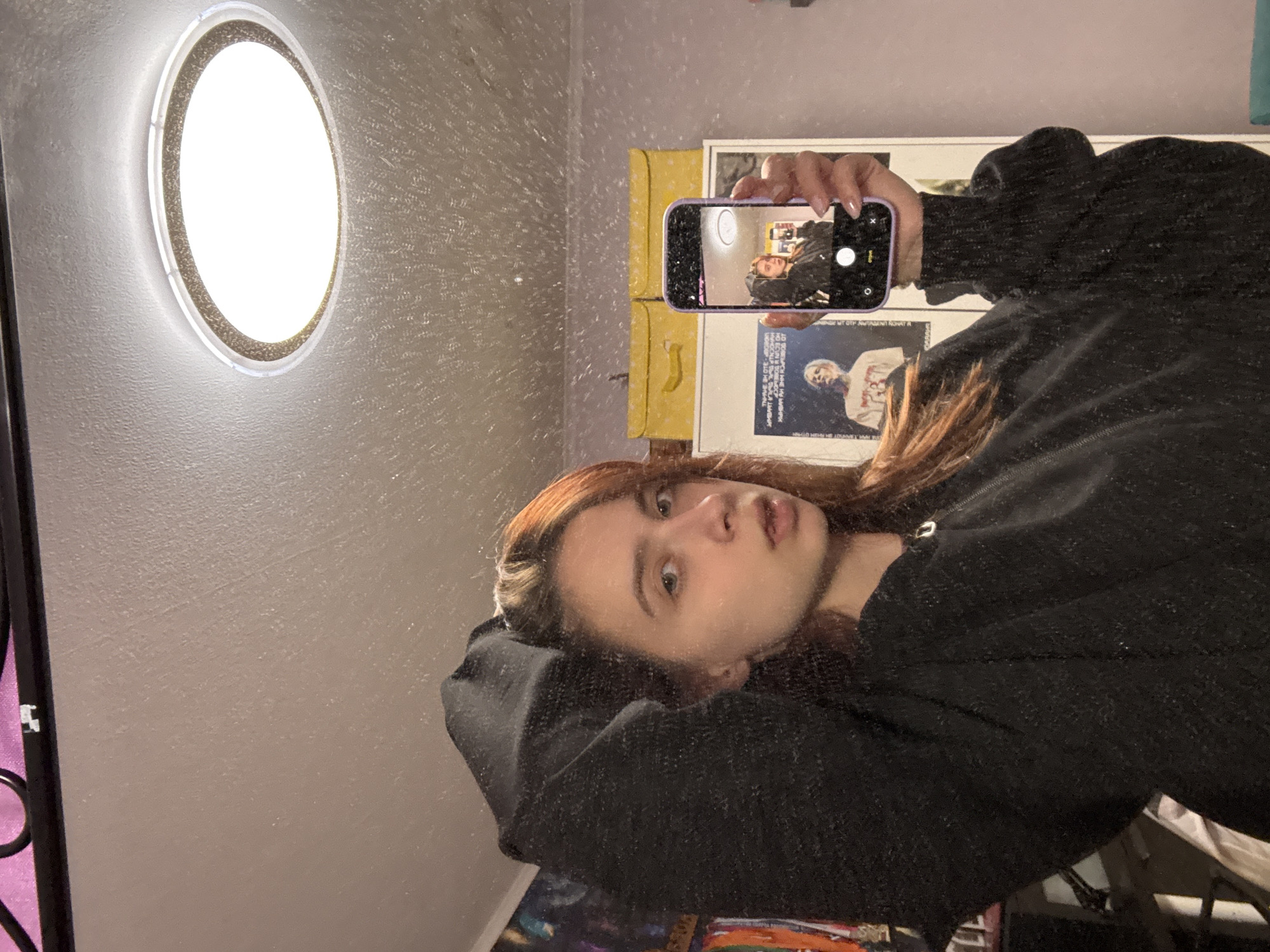 A person with auburn hair takes a mirrored selfie in what appears to be an interior space. They are wearing a dark-colored, textured garment with large sleeves. The phone in the person’s hand reflects a similar image of the person. In the background, a round, illuminated ceiling light is visible. Also present are posters or framed art hung on the wall. The location in the city of Прикамский, Russia, is not discernible from the image.