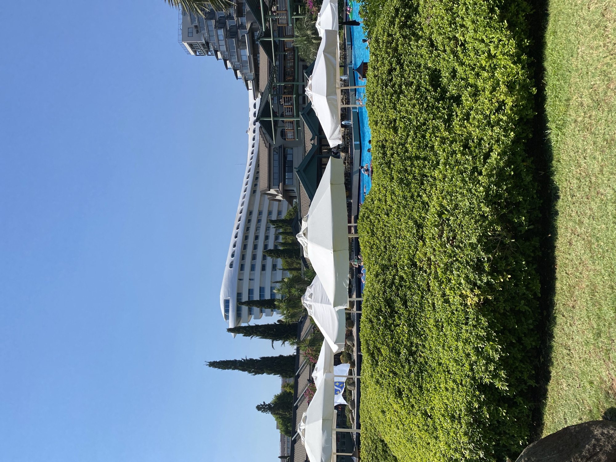 In Aksu, Turkey, a sunny day is depicted with a resort's swimming pool and surrounding landscape. In the foreground, a dense, neatly trimmed green hedge forms a visual barrier. Behind the hedge, a bright blue swimming pool is visible, with several people swimming and wading in the water. Several white beach umbrellas are set up around the pool area, providing shade. In the middle ground, a multi-story resort complex is situated, featuring dark green roofing and balconies. Further in the background, a uniquely designed white building with a curved, aerodynamic shape stands out against the clear blue sky. The architectural style of the resort suggests a modern, upscale establishment. Several tall, slender trees, likely cypress trees, are interspersed throughout the landscape, adding vertical elements to the scene. The overall impression is one of a vacation destination with ample amenities and a pleasant outdoor environment.
