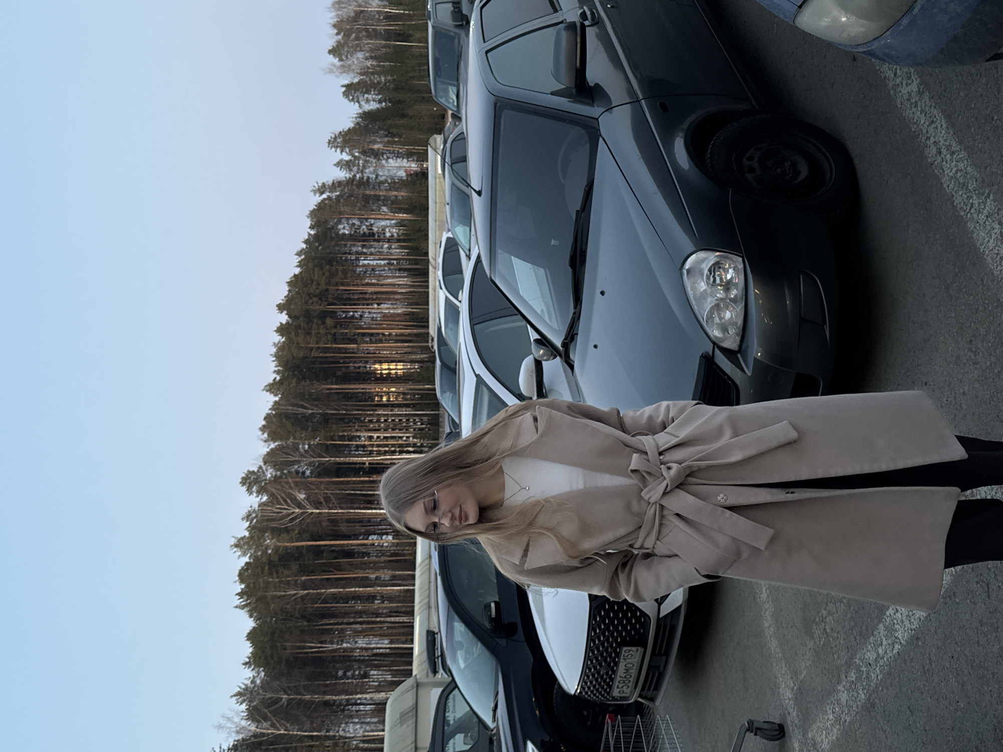 A woman stands in a parking lot in Perm, Russia. She is wearing a light beige trench coat, white top, glasses, and dark pants. The woman is positioned between rows of parked cars. The license plate "P586MO 159" is visible on a white car. Tall trees are in the background. The sky is light.