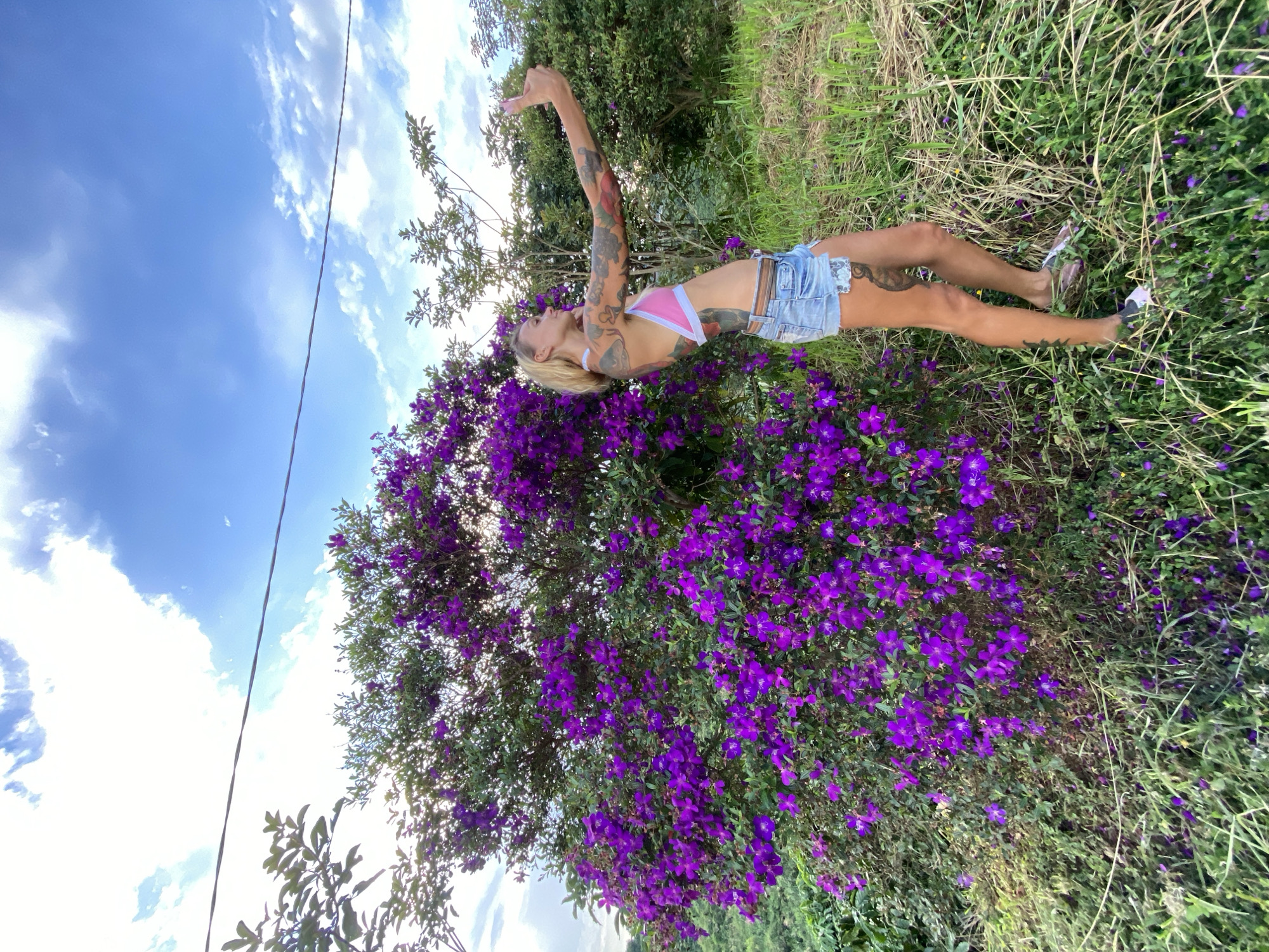 A Caucasian woman with blond hair and extensive tattoos on her arms and torso stands to the right of a large bush with vibrant purple flowers. She is wearing a pink and white bikini top and denim shorts, and she is holding up a smartphone to take a selfie. The woman is positioned on a grassy slope. Behind her, there are more trees and foliage. Above, a bright blue sky is visible with scattered white clouds, and a thin black wire stretches horizontally across the upper portion of the image. The scene appears to be outdoors in a rural or natural setting, not specifically identified within the city of Thôn 3, Vietnam.