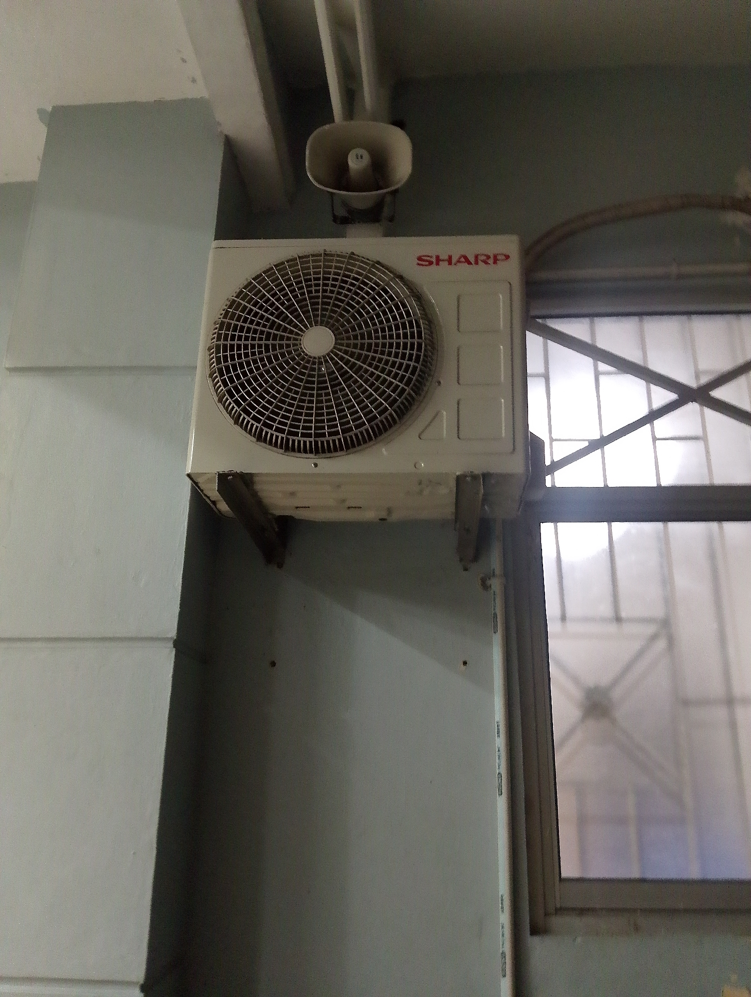An outdoor unit of an air conditioner is mounted on a light blue wall. The air conditioner unit is white and has a circular grille with a fan behind it. The brand name "SHARP" is visible in red lettering on the unit. Above the air conditioner, a white horn speaker is mounted. To the right of the air conditioner, there is a window with a metal grille and frosted glass. The scene appears to be in an outdoor or semi-outdoor area, possibly a corridor or a building exterior. The image is taken from a slightly low angle, looking up towards the air conditioner and speaker. The lighting suggests it might be daytime, with natural light coming through the window. The wall has a slightly textured finish. There are no people visible in the image. The overall condition of the equipment appears functional but shows signs of exposure to the elements. This type of installation is common in urban environments in Southeast Asia, including in the city of Pematang Siantar, Indonesia, where buildings often feature outdoor air conditioning units mounted on walls for cooling purposes, especially in residential and commercial establishments. The horn speaker suggests a public address system, which could be for announcements or alarms.