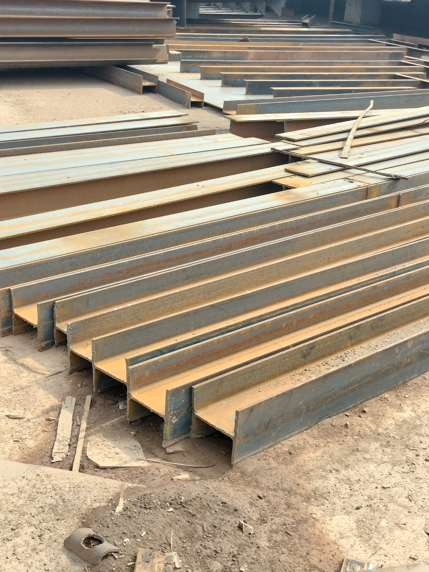 A construction material storage yard in Batam, Indonesia, features rows of weathered I-beams stacked and laid out on uneven, dusty ground. The I-beams, some showing signs of rust and oxidation, are arranged parallel to each other, creating a strong visual pattern. Scattered on the ground are smaller pieces of lumber and other debris. The lighting suggests an outdoor environment under daylight. The scene is devoid of people.