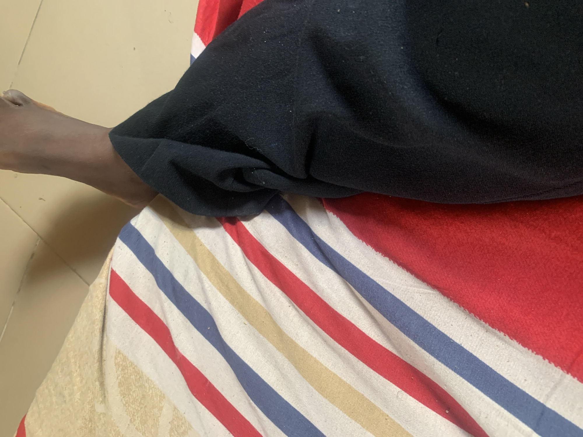 In Jauro Yinu, Nigeria, a close-up shot shows a person's bare foot and lower leg, clad in black pants, resting on a striped blanket. The blanket features diagonal stripes of white, red, blue, and tan. The background consists of a light-colored, tiled floor.