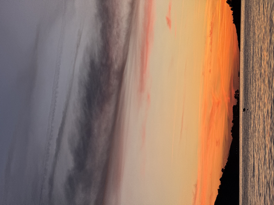 A landscape image shows a colorful sunset scene. To the left, there is a gray and blue sky with wispy clouds. The sky transitions to orange and yellow hues on the right, with hints of pink. A dark, silhouetted landmass appears along the right side, with water reflecting the sunset's warm colors. The overall composition emphasizes the sky's gradations and the tranquil atmosphere of the sunset.