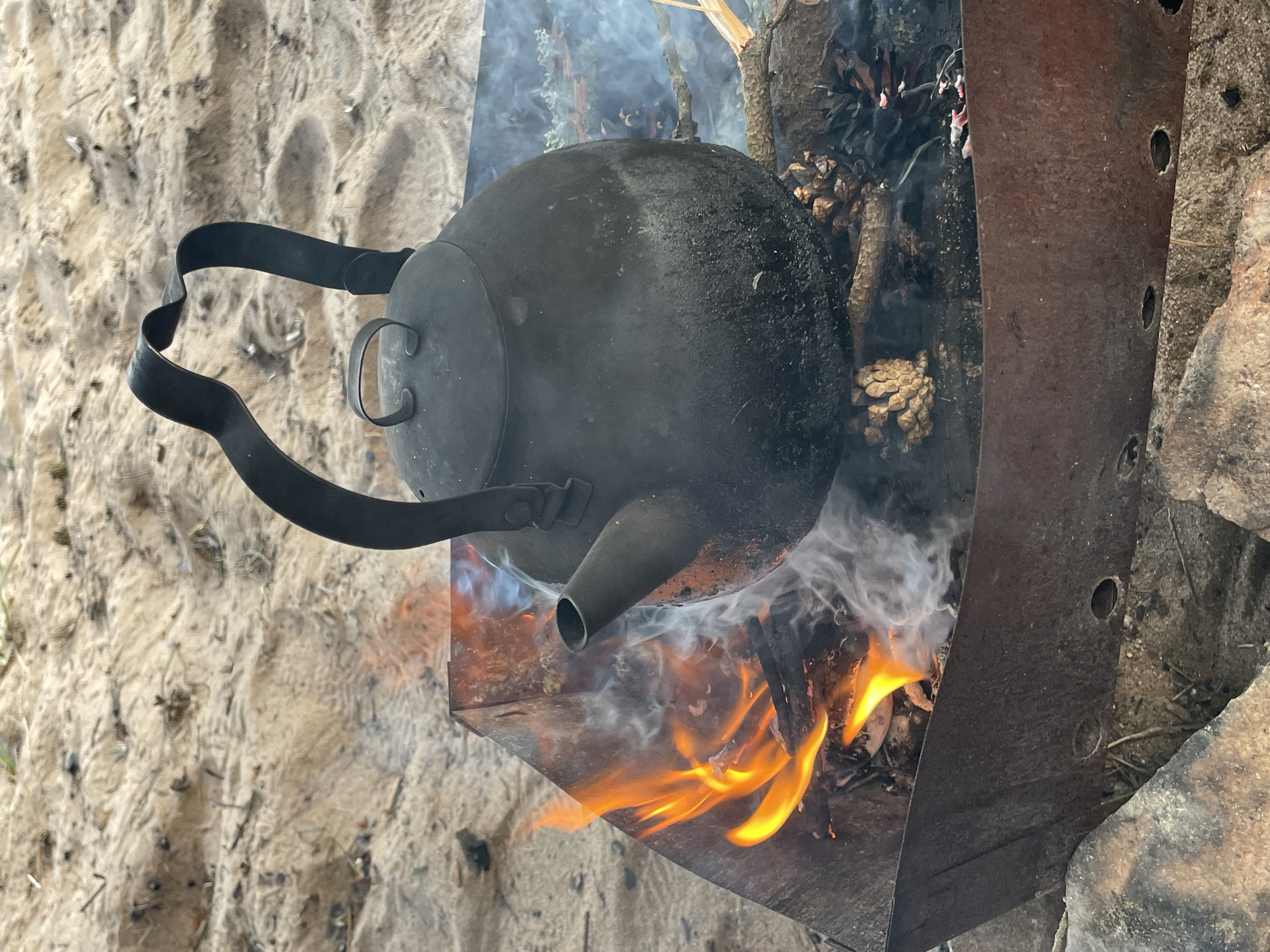 A close-up, vertical shot captures a dark, rustic kettle suspended over a campfire on a sandy surface. The kettle, made of a dark metal, rests on charred wood and pinecones within a weathered metal fire pit. Flames lick up from the burning wood, producing wisps of white smoke that curl around the kettle. The background is a sandy beach with sparse vegetation. The image appears to be taken outdoors, possibly in a natural setting. The location is specified as Unknown, Russia. There are no people or animals visible in the image, and no discernible interactions are occurring beyond the kettle being heated by the fire.