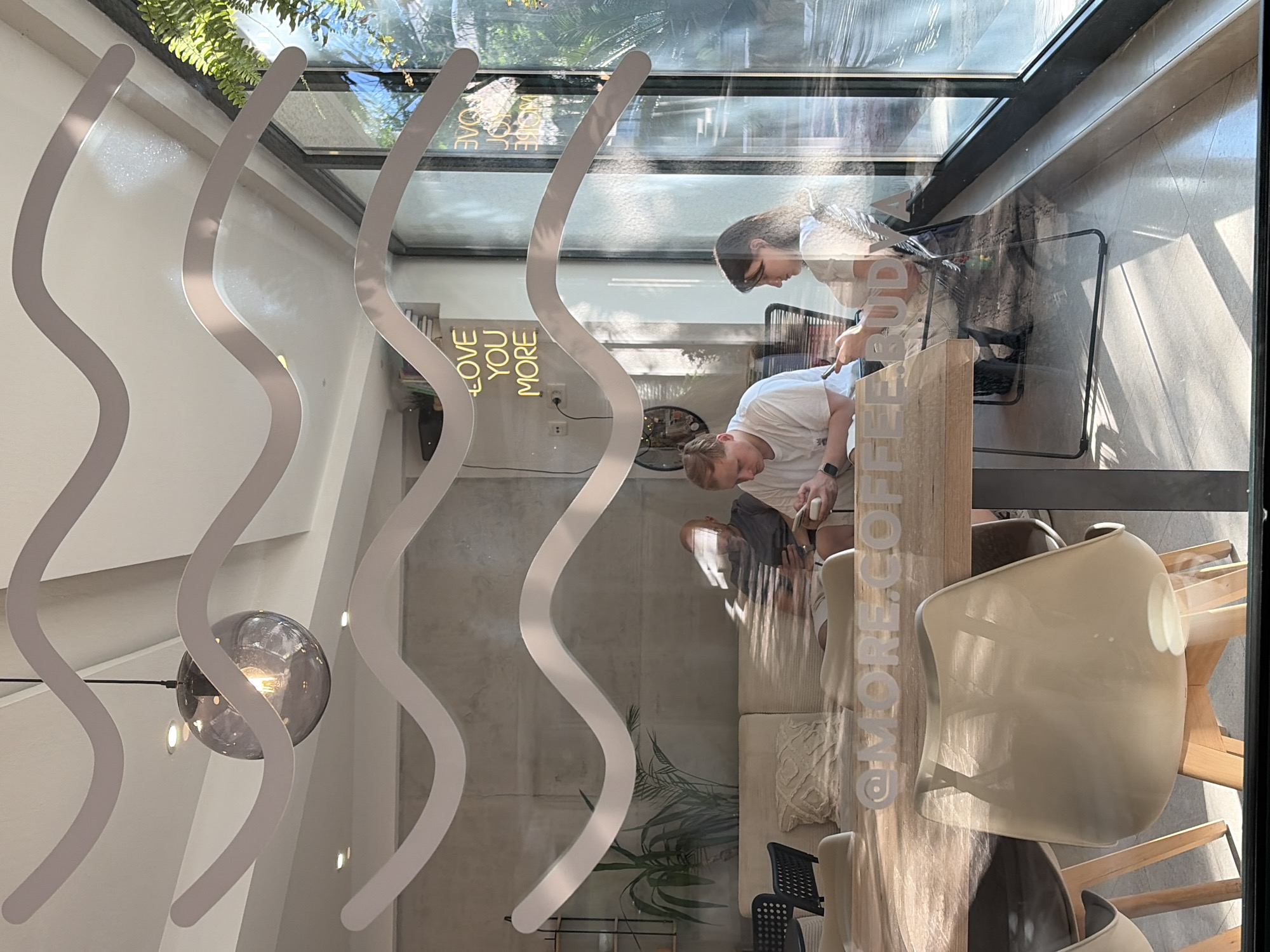 The image depicts the interior of a modern coffee shop located in Budva, Montenegro, as indicated by the reflection of a neon sign that reads "LOVE YOU MORE" and "BUDVA". The perspective is from outside the coffee shop, looking through a window adorned with horizontal white wavy lines.
Inside, a man with short blonde hair wearing a white t-shirt is seated at a wooden table, looking down at his mobile phone. To his right, a woman with dark hair, also wearing a white t-shirt, is seated and looking towards her own phone. In the background, a man with a shaved head, wearing a dark t-shirt, is seated on a light-colored couch and is also looking at his phone. A potted plant is visible to the left of the couch. The walls appear to be made of concrete.
The foreground features a wooden table with chairs around it. The chairs are made of a light-colored molded plastic with wooden legs. Sunlight streams in through the large windows, casting shadows on the floor and furniture. The overall ambiance is casual and relaxed, suggesting a typical daytime scene in a coffee shop.