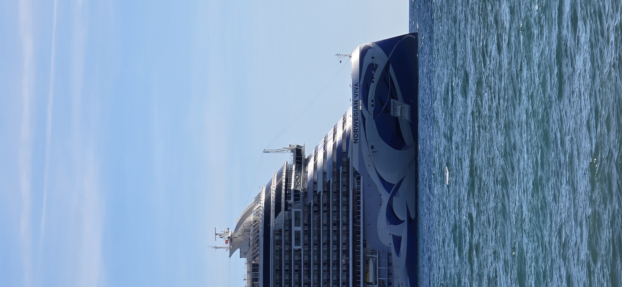 A large cruise ship, the Norwegian Viva, is docked at sea, possibly near Bar, Montenegro. The ship is predominantly white with blue wave-like designs on its hull and the text "NORWEGIAN VIVA" clearly visible. The ship's structure features multiple decks with rows of windows, a prominent bridge, and various antennae and navigational equipment on its upper levels. The ship is positioned against a backdrop of a clear blue sky with a few wispy white clouds. The foreground is occupied by choppy, blue ocean water.