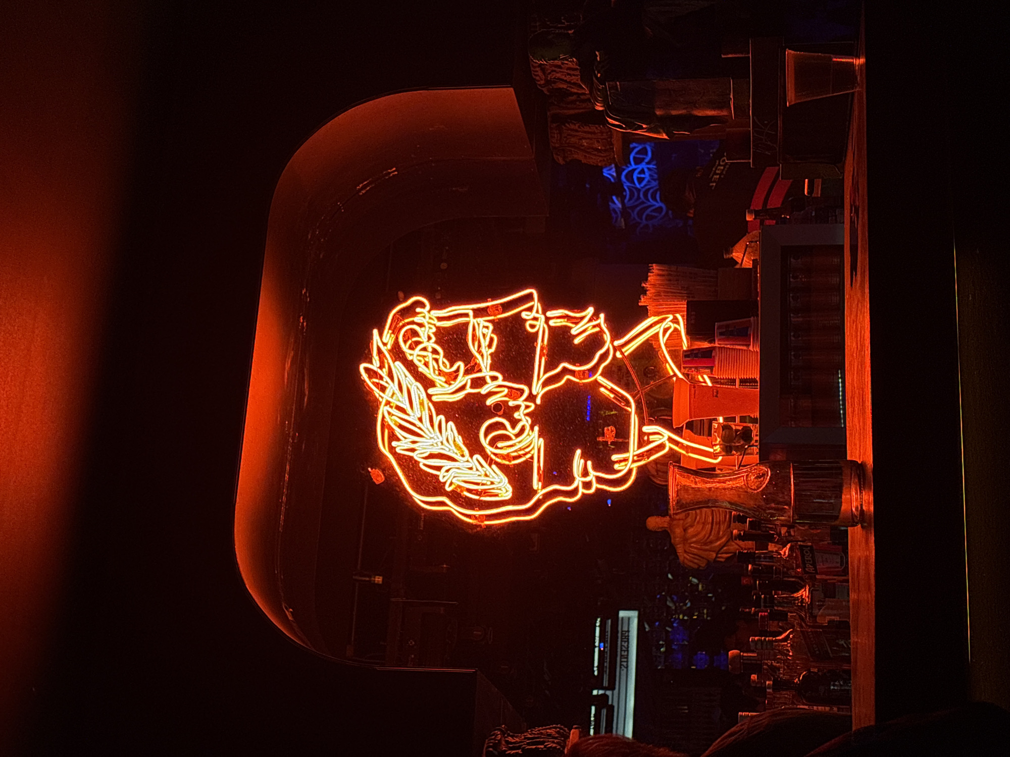 The image displays an interior scene within a bar or club in San Bernardino Tlaxcalancingo, Mexico, under dark, predominantly red lighting conditions. A prominent, bright red and yellow neon sign in the upper center depicts a stylized profile bust of a male figure, resembling a Roman or Greek emperor, adorned with a laurel wreath on his head.
Below the neon sign, a dark, reflective bar counter is visible. On the left side of the counter, numerous liquor bottles, including identifiable labels such as "Red Label" and "Aperol," are arranged. Adjacent to these are a clear glass carafe partially filled with liquid and an empty clear drinking glass. To the right of the carafe, a stack of white plastic cups and a cluster of drinking straws are present. A clear plastic cup containing a reddish liquid rests on the far right of the counter. The extreme lower-left foreground shows a dark, indistinct silhouette, consistent with a person's shoulder and head.
The background features a dark, arched architectural element above and behind the neon sign, reflecting some of the red light. Various dark, classical-style statues or busts are discernable as decorative elements behind the bar. Faint blue light patterns and reversed text, reading "ZЯƎBUI2," are visible in the deeper background, suggesting additional signage or decorative lighting. Mounted speakers are also visible on the upper left wall.
