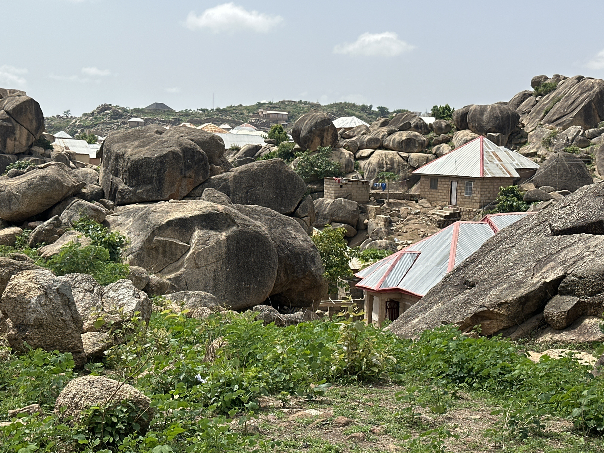 The image depicts a rocky landscape in the city of Jos, Nigeria, characterized by large granite boulders interspersed with vegetation and scattered residential buildings. The buildings are modest, with corrugated metal roofs, some painted red. The foreground is dominated by green foliage. Other homes are built into the landscape of rocks. The sky is overcast, with a few visible clouds. A town with multiple buildings and a telecommunications tower can be seen in the distance. No people are visible.