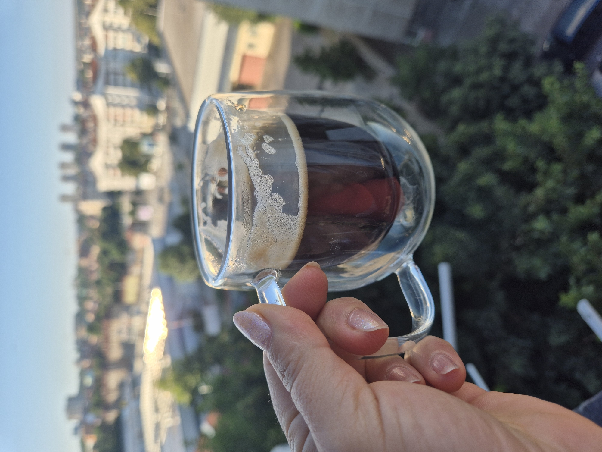 A person's hand, with manicured nails, holds a clear glass mug filled with dark coffee. The mug has a double-walled design and a small, transparent handle. The coffee has a layer of crema on top. The background is a blurred cityscape with buildings and trees, suggesting an urban environment. The lighting indicates it is daytime, possibly morning or late afternoon.