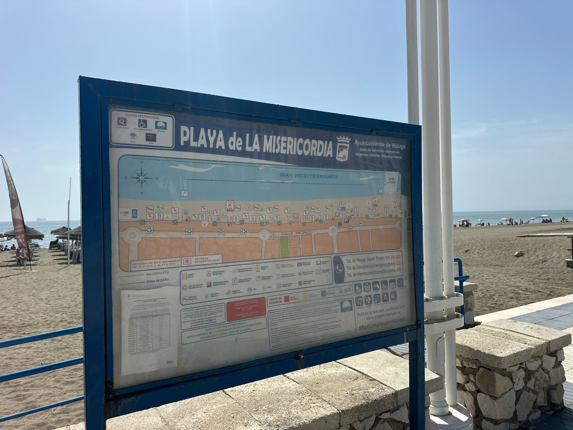The image shows a blue sign with a map of La Misericordia beach in Málaga, Spain. The sign is mounted on a metal frame and is located on a sandy beach. The map displays various facilities and services available at the beach, such as restrooms, showers, and information points. There are also icons indicating accessibility features, recycling points, and emergency services. In the background, people can be seen relaxing on the beach, with umbrellas and sun loungers. The sea is calm and the sky is clear and blue. The sign also includes information about the beach's certifications, such as ISO 9001 and the Blue Flag award. The Ayuntamiento de Málaga (City Council of Málaga) is mentioned as the authority responsible for the beach. There are also contact numbers for beach services and emergencies. The hashtag #SerLimpiosporMálaga is visible at the bottom of the sign.