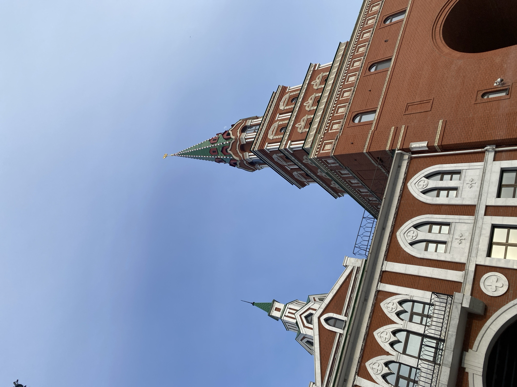 The image captures an upward-looking perspective of two architecturally distinctive buildings against a clear, bright blue sky, located in Yoshkar-Ola, Russia.
The most prominent feature is a tall, multi-tiered tower on the left side of the frame, constructed from red brick with elaborate white and green decorative stonework. This tower features a stepped design, oval windows, and culminates in a sharp, green-tipped spire topped with a golden finial. Its design evokes historical Kremlin-style architecture.
In the foreground and to the right, another building of similar red brick and white decorative elements is visible. This structure boasts a facade with multiple arched windows, some featuring intricate tracery, and at least two balconies with dark iron railings. A smaller, green-tipped spire is also visible on a pitched roof section of this building, aligning with the architectural style of the taller tower.
The clear, cloudless blue sky indicates fair weather and bright daylight conditions. No people or specific activities are depicted, allowing the focus to remain on the unique architectural details of the structures. The buildings showcase a blend of styles, with ornate brickwork and stone accents, characteristic of the deliberately designed urban landscape found in Yoshkar-Ola. There is no visible text in the image.