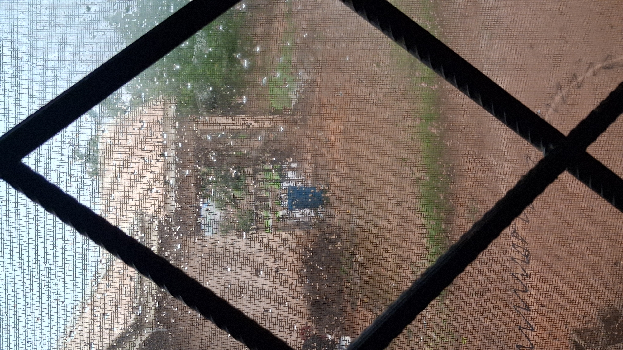 The image is taken from inside a building in Barakin Akawo, Nigeria, looking through a screen and a diamond-shaped metal grate. The view outside shows a rural or semi-rural environment with a dusty, reddish-brown ground and some buildings with tiled roofs. It appears to be raining, as evidenced by the numerous water droplets on the screen and the generally wet appearance of the ground and foliage. There is a blue barrel or container visible in the middle ground, as well as some fencing and vegetation. The overall atmosphere is subdued, likely due to the overcast weather. The screen itself has a fine mesh, and there are some indistinct markings or scribbles on its surface, particularly in the lower left quadrant. The metal grate is black and has a twisted or coiled texture.