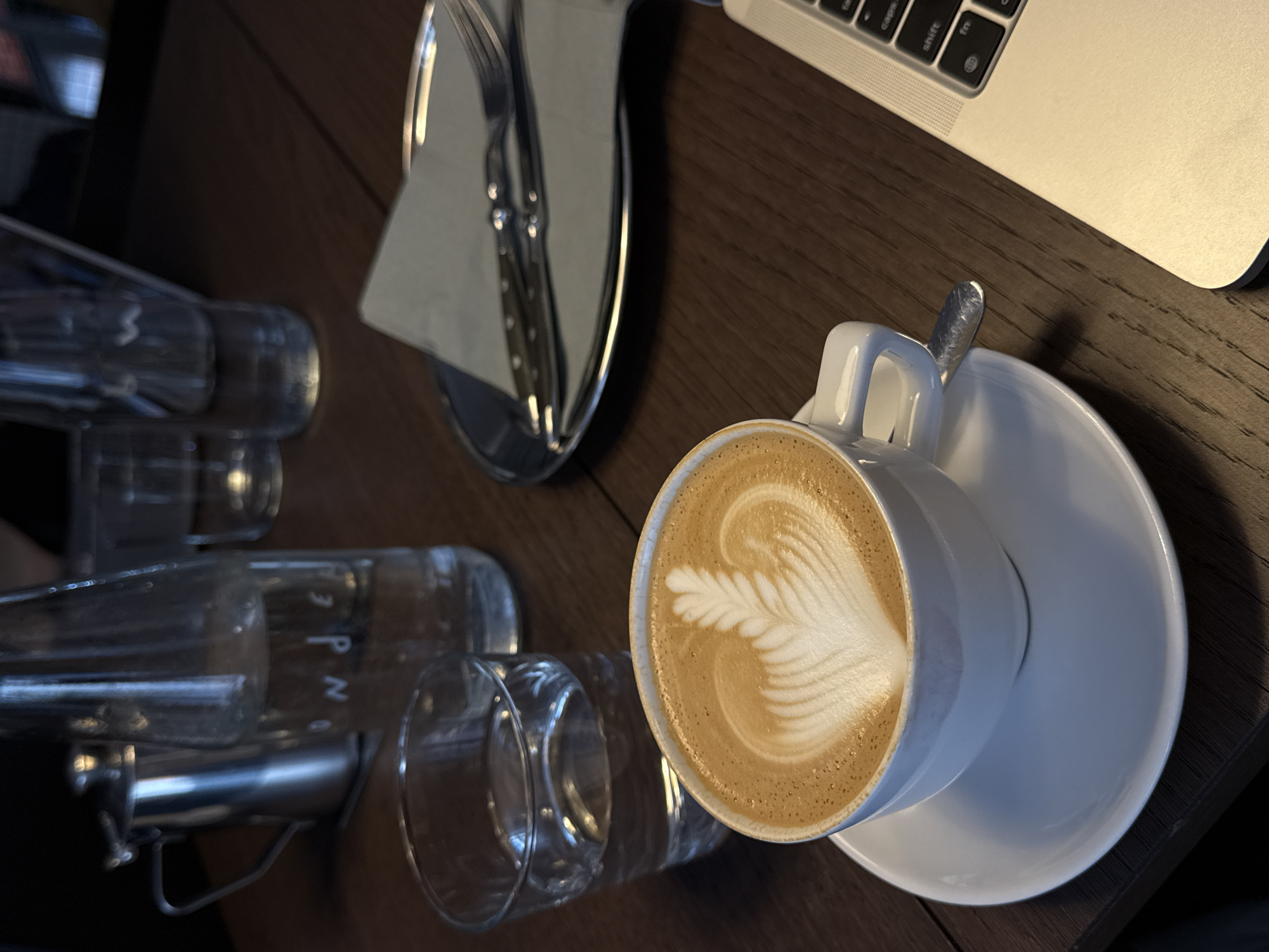 The image depicts a wooden table in what appears to be a cafe or restaurant setting. A cup of coffee with latte art sits on a white saucer, with a spoon placed on the saucer. Next to the coffee are several empty glasses in a metal frame, and cutlery in a silver tray. A laptop is partially visible in the upper right corner. The location is suggested to be Belgrade, Serbia. There are no people visible in the image. The lighting is warm.