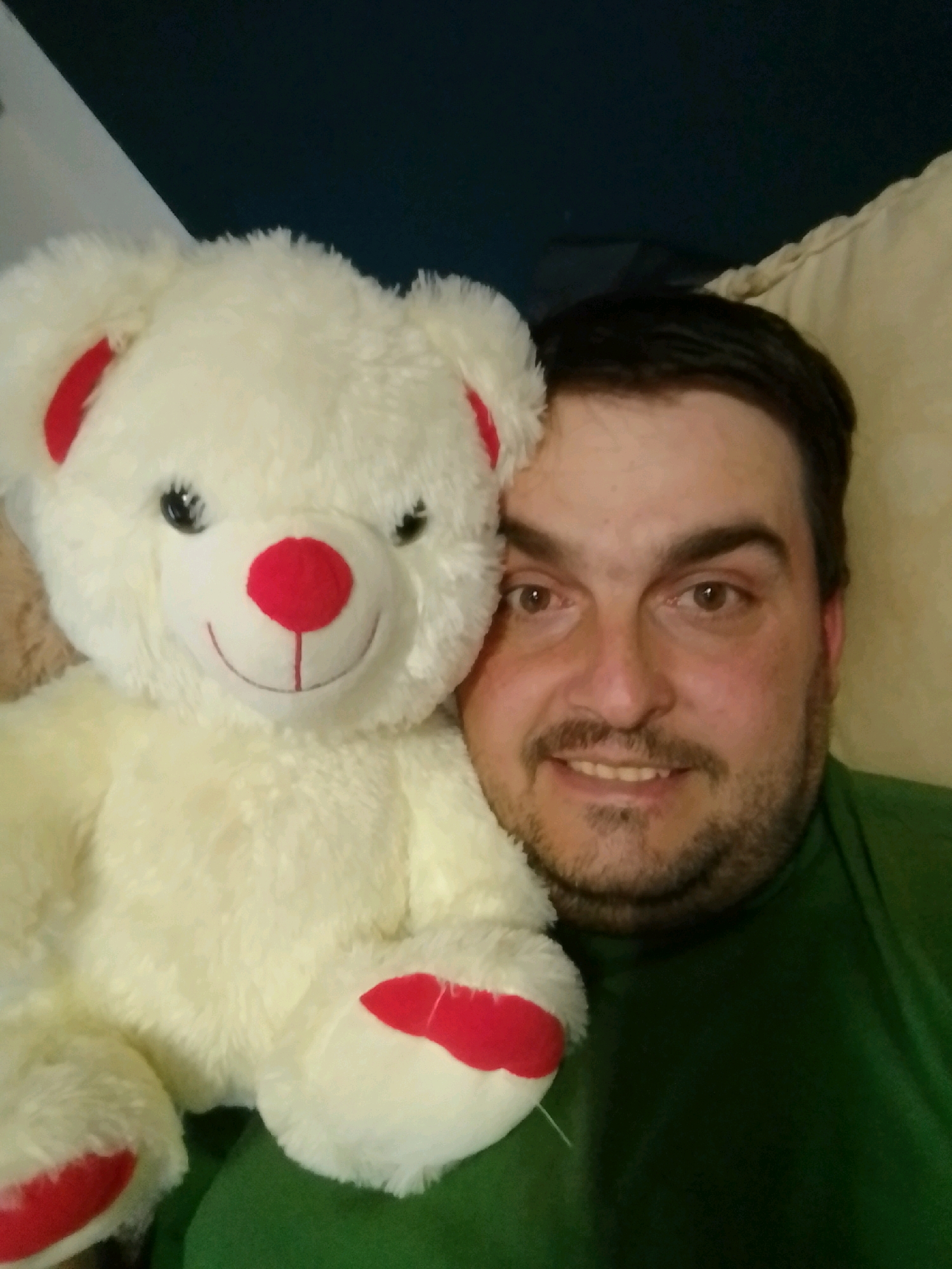 The image shows a close-up, overhead, selfie-style shot of a man and a white teddy bear. The man has dark hair, brown eyes, and a beard with a mustache. He is wearing a green t-shirt. The teddy bear is positioned to his left and is partially obscuring his face. It is a white teddy bear with red accents on its ears, nose, and paws. The background appears to be indoors with a dark wall or curtain behind the man and a beige upholstered surface, possibly a couch or chair, to his right. There is no identifiable information that would allow for a specific location in the city of Wedel, Germany.