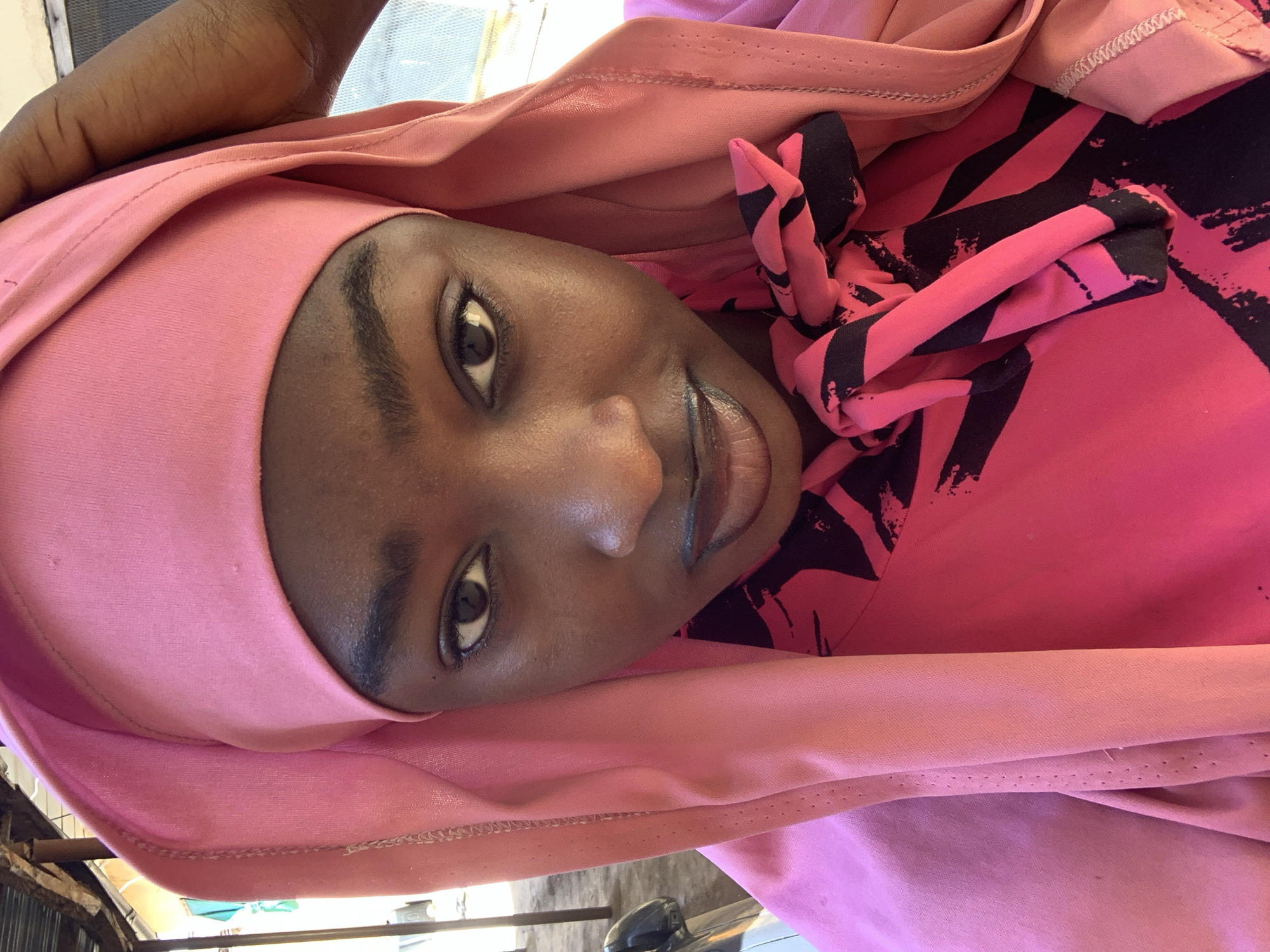 A close-up shot focuses on a woman with dark skin, dark eyes, and a pink hijab. She is wearing a pink top with a black pattern. The location is Rafin Par, Nigeria. The background shows parts of a building and vehicle.