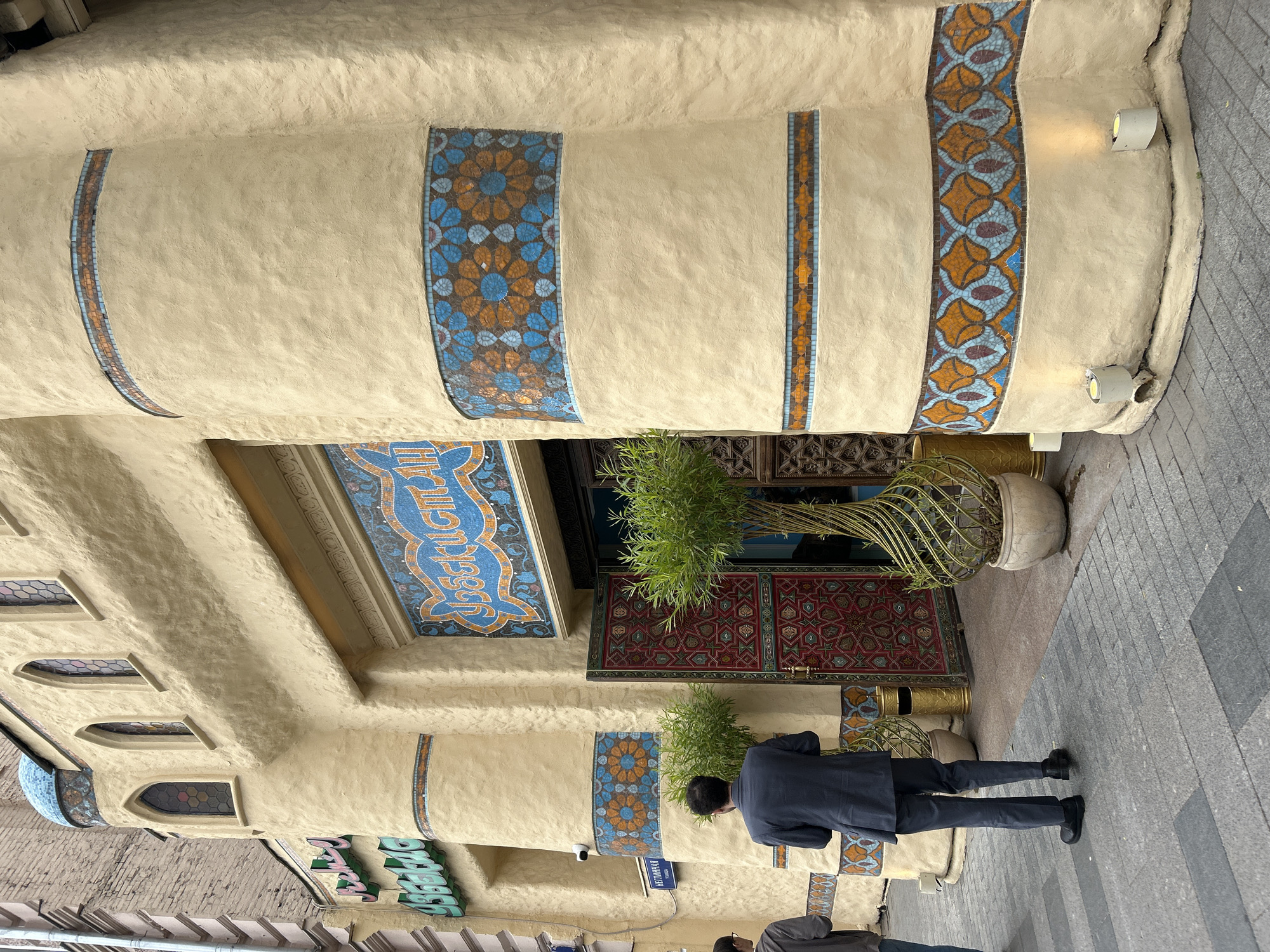 The image shows the entrance of a building in Moscow, Russia, with a beige, textured facade decorated with colorful mosaic tiles in blue and orange floral patterns. Above the entrance, a sign in blue and orange script reads "Узбекистан" (Uzbekistan). The entrance doors are ornate, with intricate geometric patterns in red and green. Two people are visible on the sidewalk in front of the building; one man in a dark suit and another person in a grey hoodie. Two potted plants with thin, twisted trunks and green foliage flank the entrance. The ground is paved with grey interlocking bricks. The building appears to be a restaurant or establishment with an Uzbek theme. A street sign that reads "Неглинная улица" (Neglinnaya Street) is visible on the wall to the left.