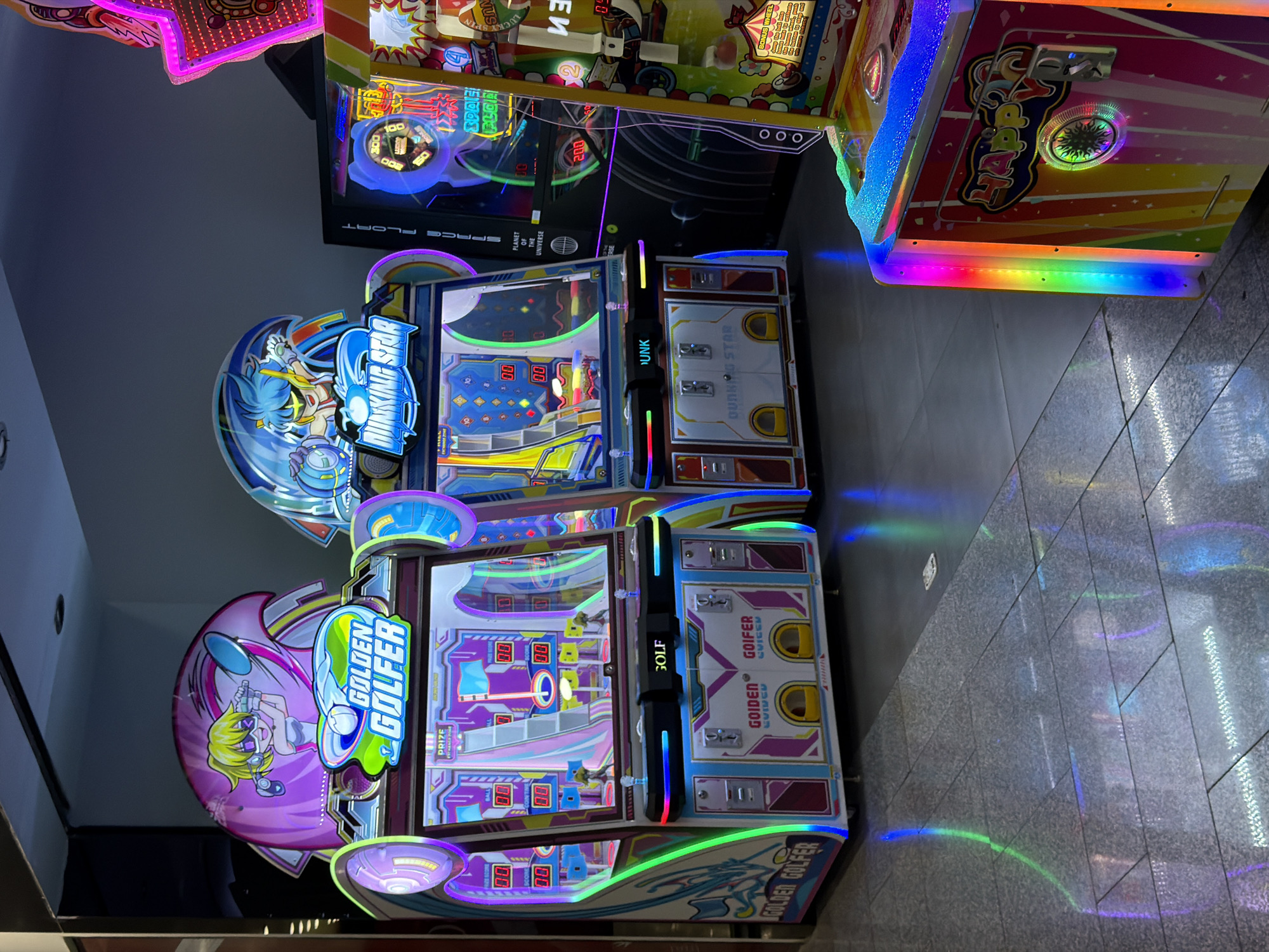 The media file shows a collection of arcade games in what appears to be an indoor entertainment venue, possibly a mall or arcade, in Đà Nẵng, Vietnam. The scene is dominated by brightly lit, colorful arcade machines.
The prominent subjects are two arcade game cabinets. The one in the foreground is titled "GOLDEN GOLFER," featuring a cartoon character with blonde hair and a golf club. The game cabinet itself is decorated with vibrant pink, blue, and green graphics, and it has a screen displaying what looks like a golf game with scores and icons. The text "GOLDEN GOLFER" is clearly visible on the cabinet.
Behind it, another arcade game is visible, labeled "DINKING STAR." This machine also has colorful graphics and a display screen. Text such as "SPACE FLOAT," "PLANET OF THE UNIVERSE," and score indicators like "100" and "200" are present on this machine. Further in the background, more arcade games are stacked, some with neon lighting and abstract designs. One machine has the text "HAPP" and "LUCKY SPIN."
The lighting suggests an indoor environment, likely during the day or evening, as the artificial lights of the arcade games are the primary light source. The floor appears to be tiled with a dark grey material, and reflections of the arcade lights can be seen on the floor, indicating a polished surface. There are no people clearly visible in the frame, but the presence of arcade games implies people would be playing them. The overall mood conveyed by the image is one of fun and entertainment, typical of an arcade setting. The visible text includes game titles, game-related terms, and numbers.