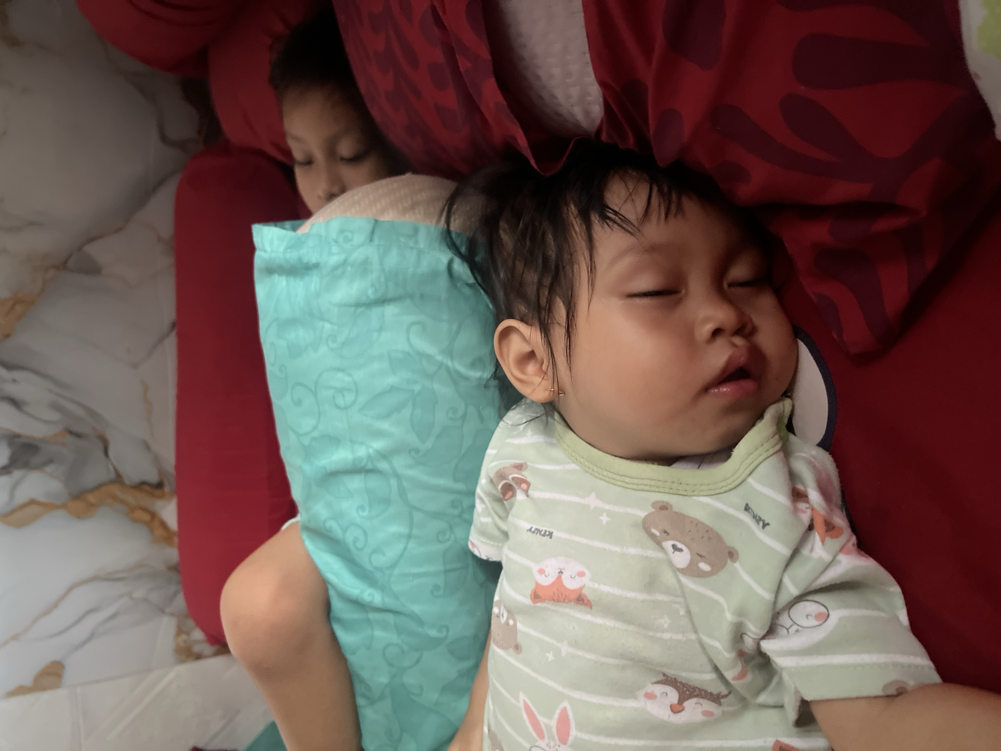 Two children are sleeping on a bed in Kecamatan Pati, Indonesia. The child in the foreground is wearing a light green onesie with white stripes and animal graphics. The child is lying on their side, facing left, with their mouth slightly open. The other child is sleeping on their back behind the first child, partially covered by a teal-colored blanket. The bed is covered with red and white bedding. The background appears to be a wall with a marble-like pattern.
