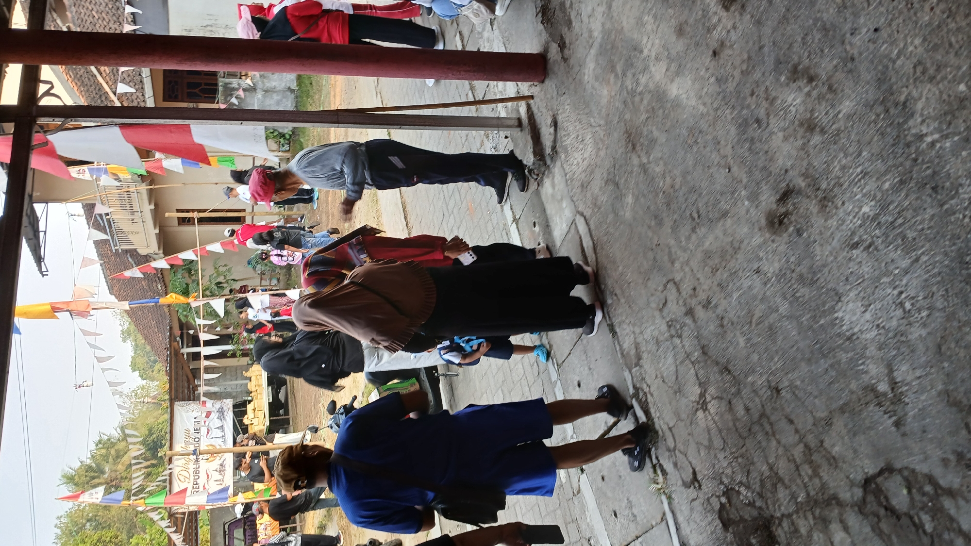 In Mendut, Indonesia, a group of people are gathered outdoors near a building adorned with Indonesian flags and pennants. The flags, in red and white, are prominently displayed along with triangular pennant banners. Several individuals are visible, including a man in a blue shirt and blue shorts, another in a grey long-sleeved shirt and dark pants, and a woman wearing a brown hijab and a long black dress. A child in a white shirt and blue shorts is also present. In the background, there is a white banner with text that appears to read "Dirga yu REPUBLIK DO IESIA," possibly related to Indonesia's Independence Day. A motorcycle is parked behind some of the people. The ground is a mix of concrete and asphalt, showing signs of wear and tear. The overall scene suggests a local celebration or event.