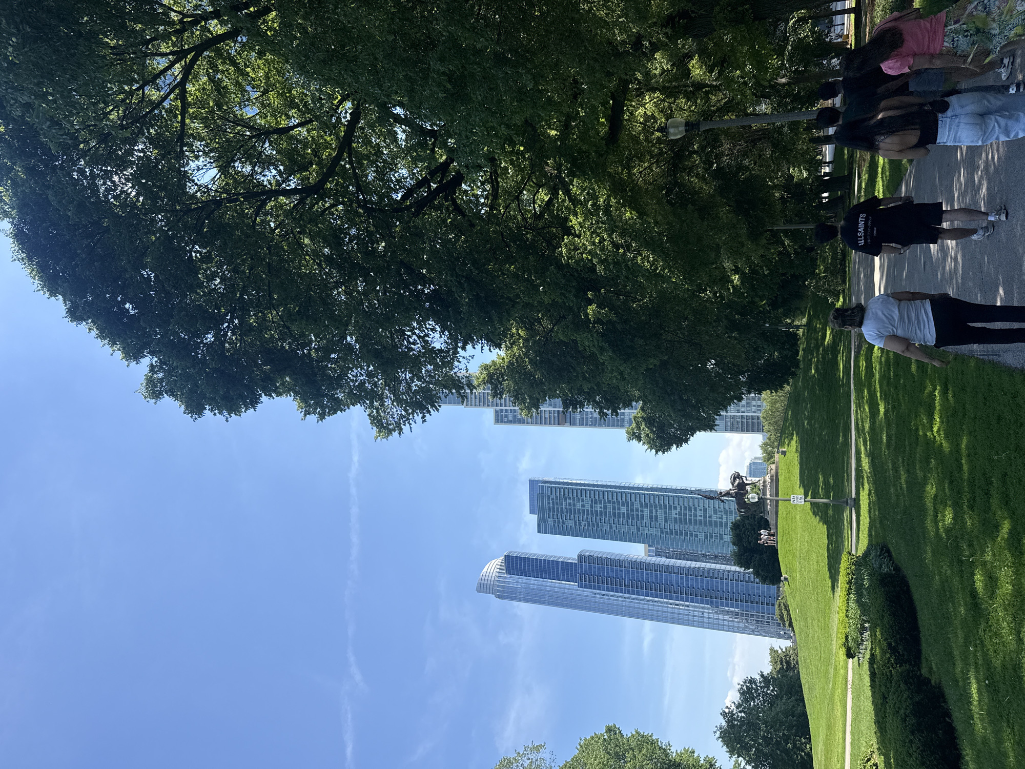 The image shows a park scene in Chicago, USA. A group of people walks along a paved path lined with green grass and trees. Skyscrapers are visible in the background, framed by the foliage of trees. A monument is visible on the grass near one of the skyscrapers. The sky is clear and blue.