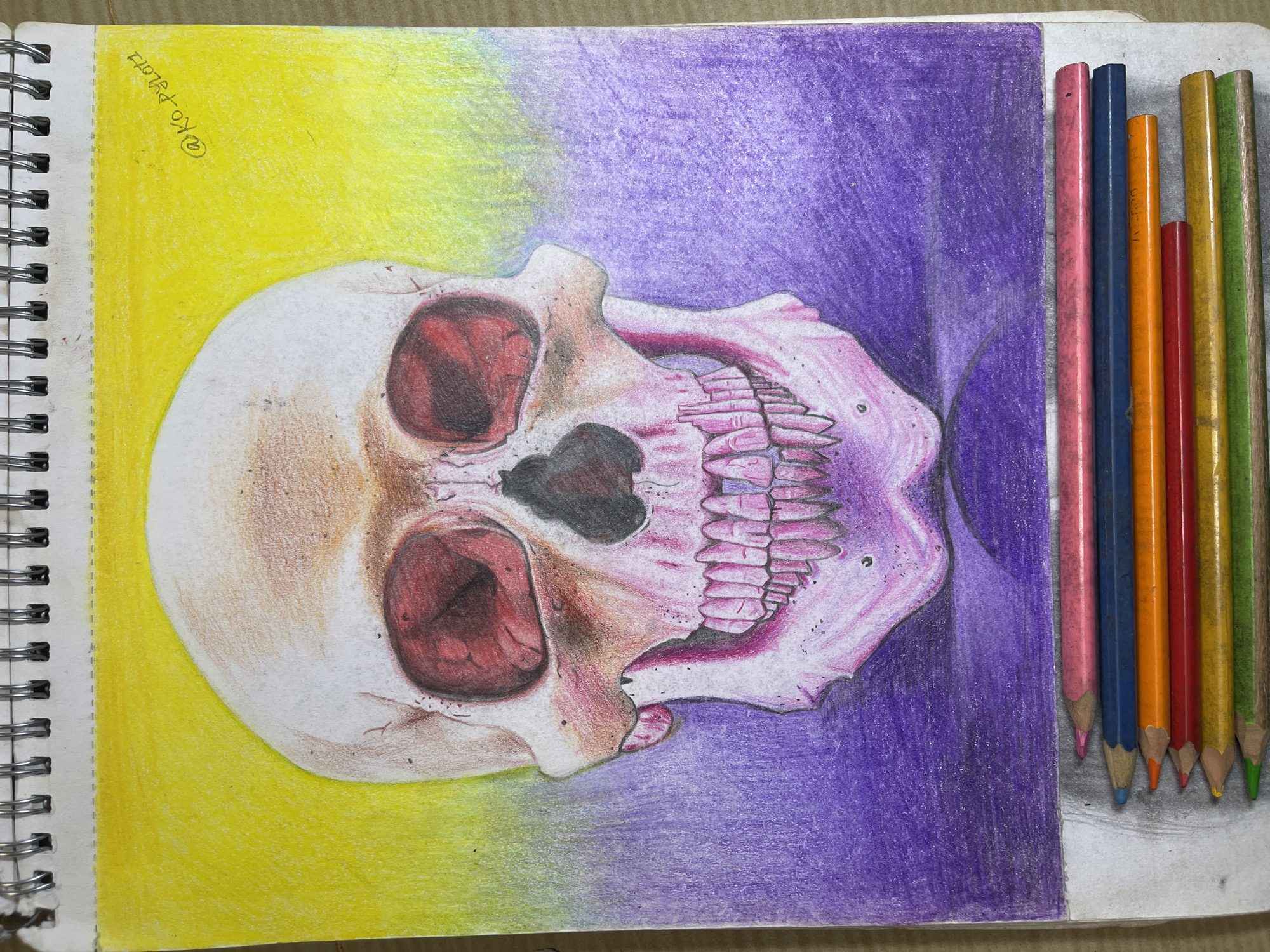 The image is a close-up, high-angle shot of a colored pencil drawing of a skull on a spiral-bound sketchbook page. The skull is rendered with detailed shading, featuring hollow eye sockets with red coloring, visible teeth, and a hint of cracked bone texture. The background of the drawing transitions from a vibrant yellow at the top to a deep purple at the bottom. In the foreground, at the bottom of the image, a set of colored pencils are laid out horizontally. The pencils are in various colors, including pink, blue, orange, red, yellow, and green. The sketchbook page appears to be resting on a textured surface, possibly a wooden table or desk. There is a handwritten signature in the upper right corner of the drawing that reads "@KO.PYLOT1". The image does not contain any elements that would suggest a location in the city of Jos, Nigeria.