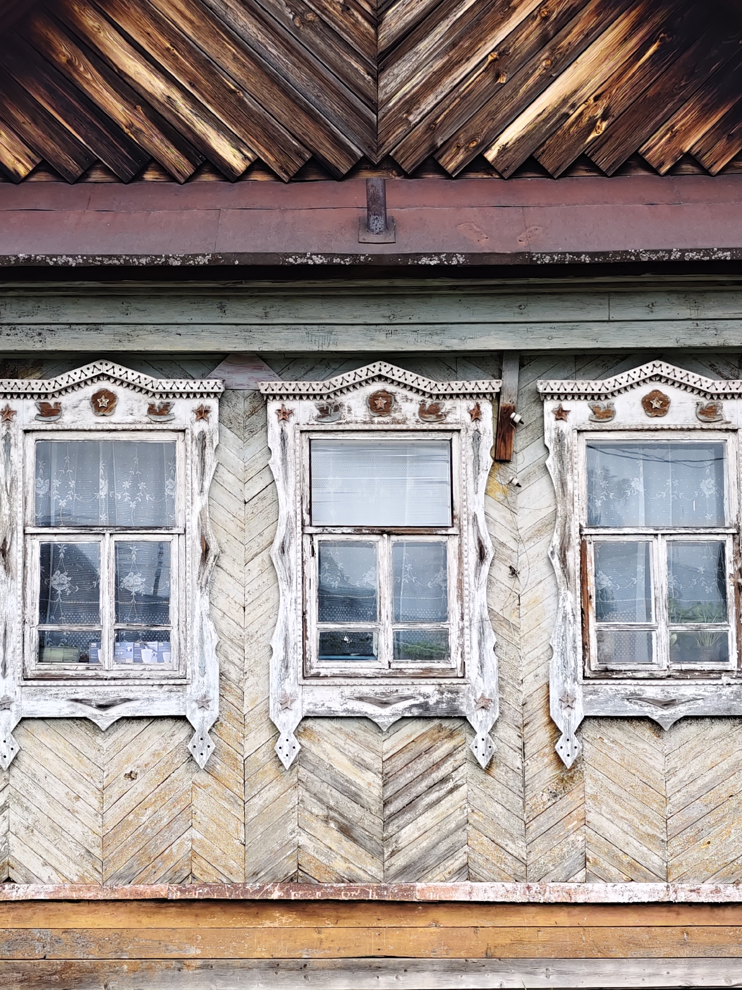 This is a close-up, vertical shot of the weathered facade of an old wooden house, characteristic of traditional Russian rural architecture. The image focuses on the upper section and three windows.
At the top, a section of the building, possibly a gable or roof line, features dark, heavily weathered wooden planks arranged in a diagonal pattern, forming a chevron. Below this, a rusted red-brown metal strip runs horizontally, with a wooden board underneath showing patches of light-colored lichen or moss.
The main wall section immediately below this consists of horizontal wooden planks, painted a faded greenish-blue, showing significant wear. Below these, the wall is covered with lighter, extensively weathered wooden planks arranged in a distinct chevron pattern. Three rectangular windows are set into this wall. Each window is adorned with elaborate, hand-carved wooden surrounds, known as "nalichniki," which are painted white but are now peeling and distressed. The carvings include intricate geometric designs, stars, and bird motifs above and beside the window panes. Behind the multi-pane glass, delicate white lace or patterned curtains are visible, and reflections suggest bright daylight.
Below the windows, the chevron-patterned wooden wall continues, eventually transitioning into horizontally laid, darker, and more robust wooden planks at the very bottom of the frame, likely forming the lower part of the wall or base.
The overall scene conveys a sense of age, craftsmanship, and the beauty of traditional materials exposed to the elements. No people or activities are visible, and there is no discernible text.