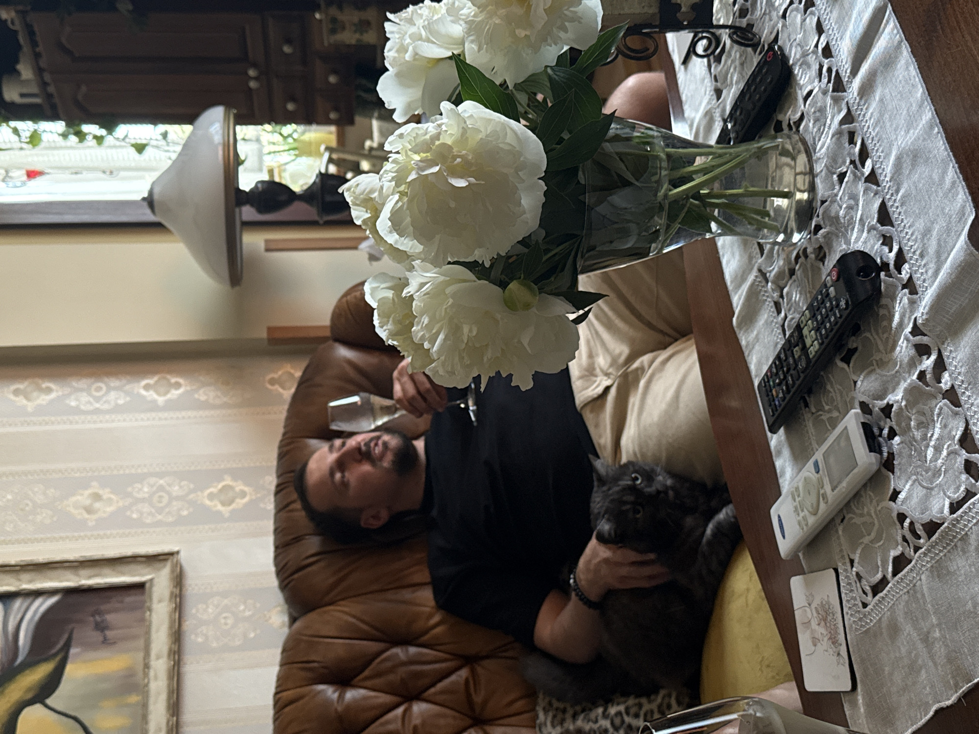 A man reclines on a brown leather couch, cradling a gray cat. He holds a wine glass in his left hand. A vase of white peonies sits on a lace tablecloth. There are two remote controls and a white device on the table. The room features patterned wallpaper and an ornate framed painting. A lamp and window are visible in the background, alongside a cabinet. The location is not discernible as being in Trnava, Slovakia, based on the provided image.