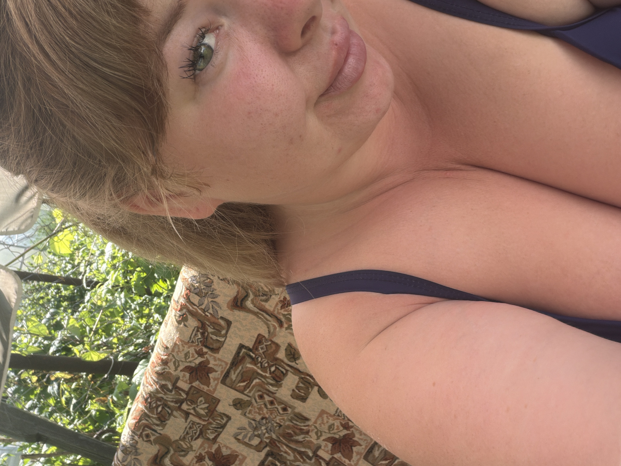 The media file contains a close-up, low-angle, portrait-style photograph of a young woman lying down outdoors. She has fair skin with freckles and light brown hair. Her eyes are green and her lips are slightly parted. She is wearing a dark blue bikini top. The background shows lush green foliage, suggesting it is daytime and likely warm weather. The woman is positioned on a patterned fabric surface, possibly a cushion or blanket, which is resting on an outdoor seating structure. The image is oriented sideways. There is no visible text. The overall mood is relaxed and casual. The location is identified as Kimry, Russia.
