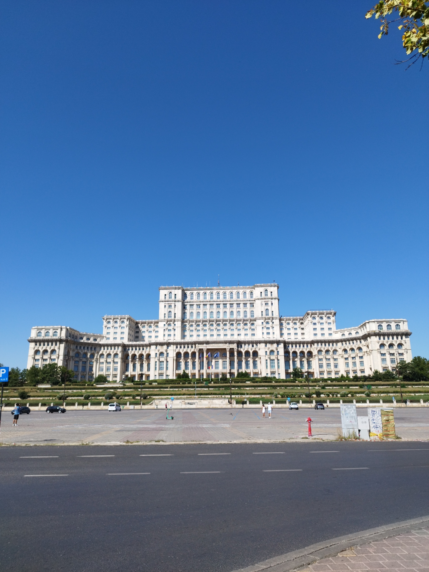 In a wide-angle shot of Bucharest, Romania, a vast expanse of clear blue sky dominates the upper portion of the frame. Below, the imposing Palace of the Parliament, a monumental neoclassical building, stretches horizontally across the middle ground. Its pale facade is adorned with numerous windows and architectural details, creating a repetitive pattern of light and shadow.
In front of the palace, a paved plaza is visible, with several vehicles parked or driving. A blue parking sign is present on the left side of the plaza. A few individuals are scattered across the plaza, some walking, others standing. A red fire hydrant stands near the right side of the plaza, adjacent to an electrical utility box covered in graffiti.
The foreground is occupied by a dark asphalt road with white lane markings. A sidewalk made of brick pavers lines the edge of the road. Greenery, including trees and manicured bushes, is present in a landscaped area between the plaza and the palace. The overall scene is bathed in bright sunlight, suggesting a clear day.