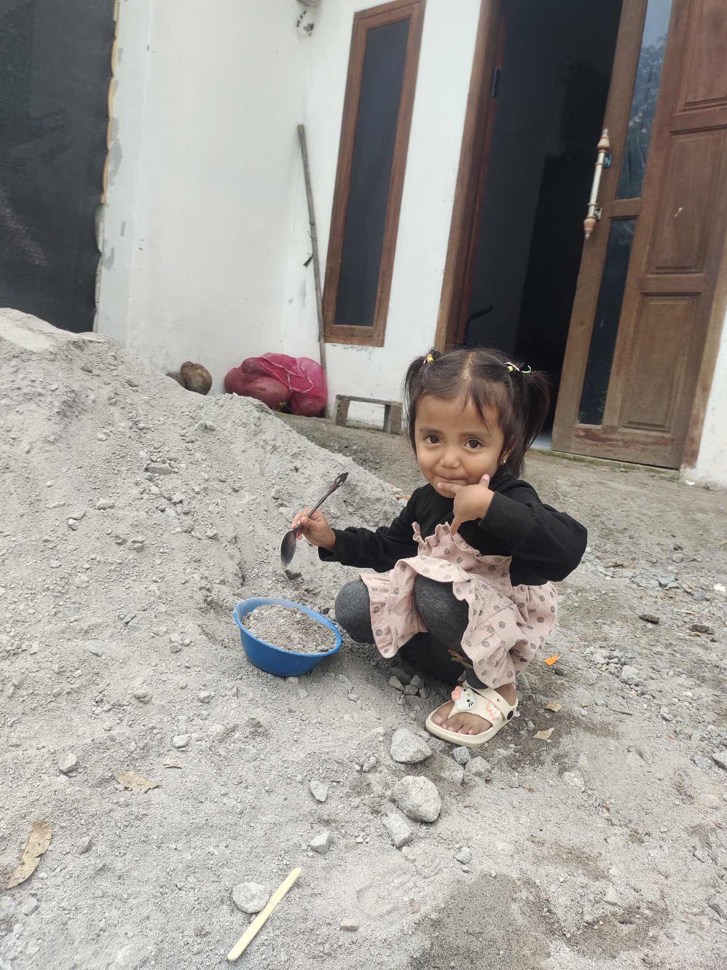 A young girl is captured squatting on a pile of sand and gravel outside a building in Cintadamai, Indonesia. The girl, wearing a black long-sleeved shirt and a light pink patterned dress over grey leggings, has her hair in two pigtails. She is holding a spoon and appears to be playing with the sand in a blue bowl. She is looking towards the camera with a slight smile and her thumb raised. The background consists of a white-walled building with wooden doors and windows, and a large pile of sand and gravel. The ground is covered with the same sand and gravel mixture. Two red mesh bags containing what appear to be root vegetables are visible on the pile of sand.