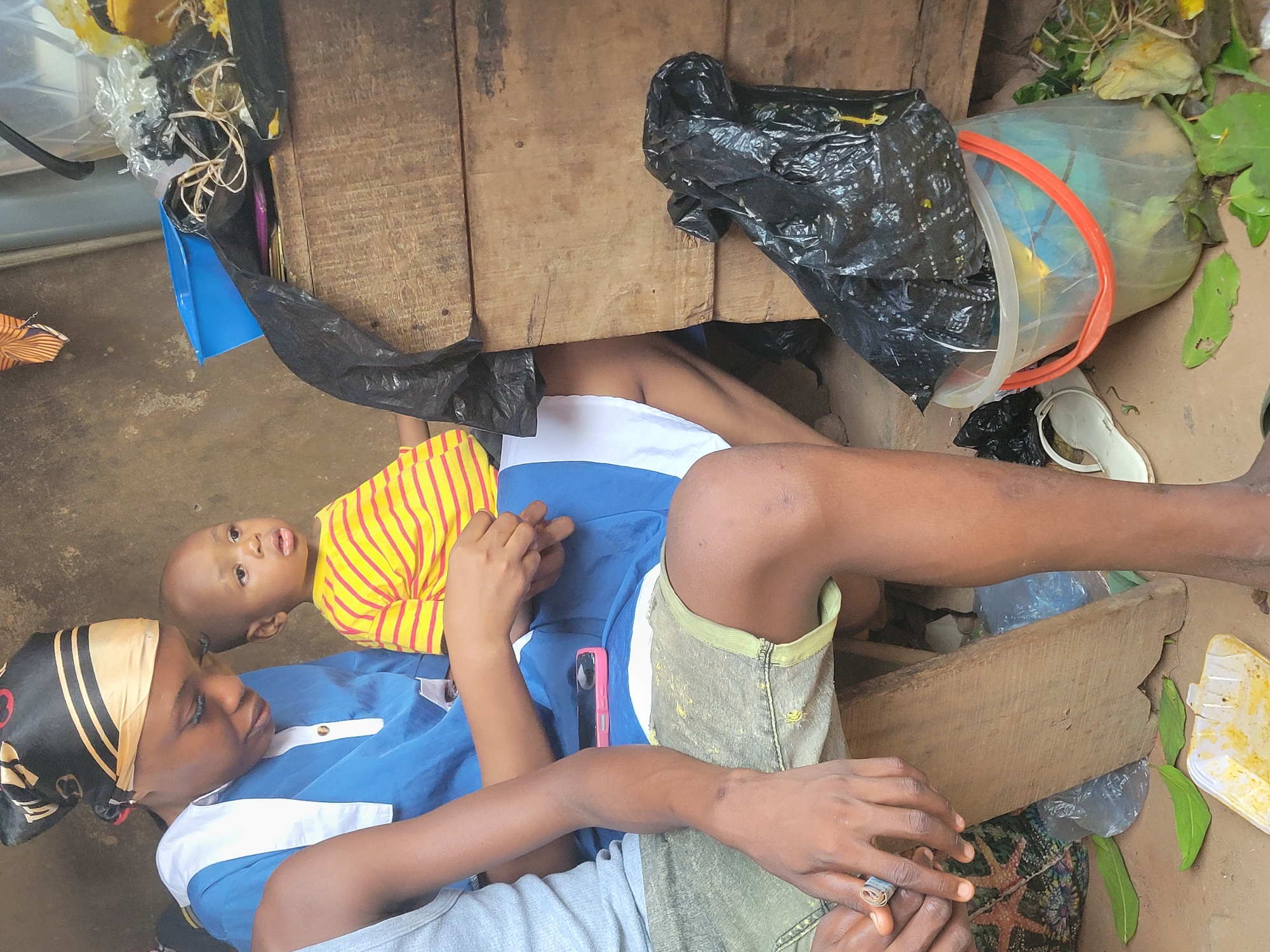 A woman wearing a blue and white top and a black headscarf is lying on the ground in Onitsha, Nigeria. She is holding a baby wearing a yellow and red striped shirt. They are lying under a wooden structure or table surrounded by debris, including a black plastic bag, a white bucket with an orange handle, a white sandal, and miscellaneous plastic containers and leaves. The ground is concrete or dirt. The scene appears to be outdoors.